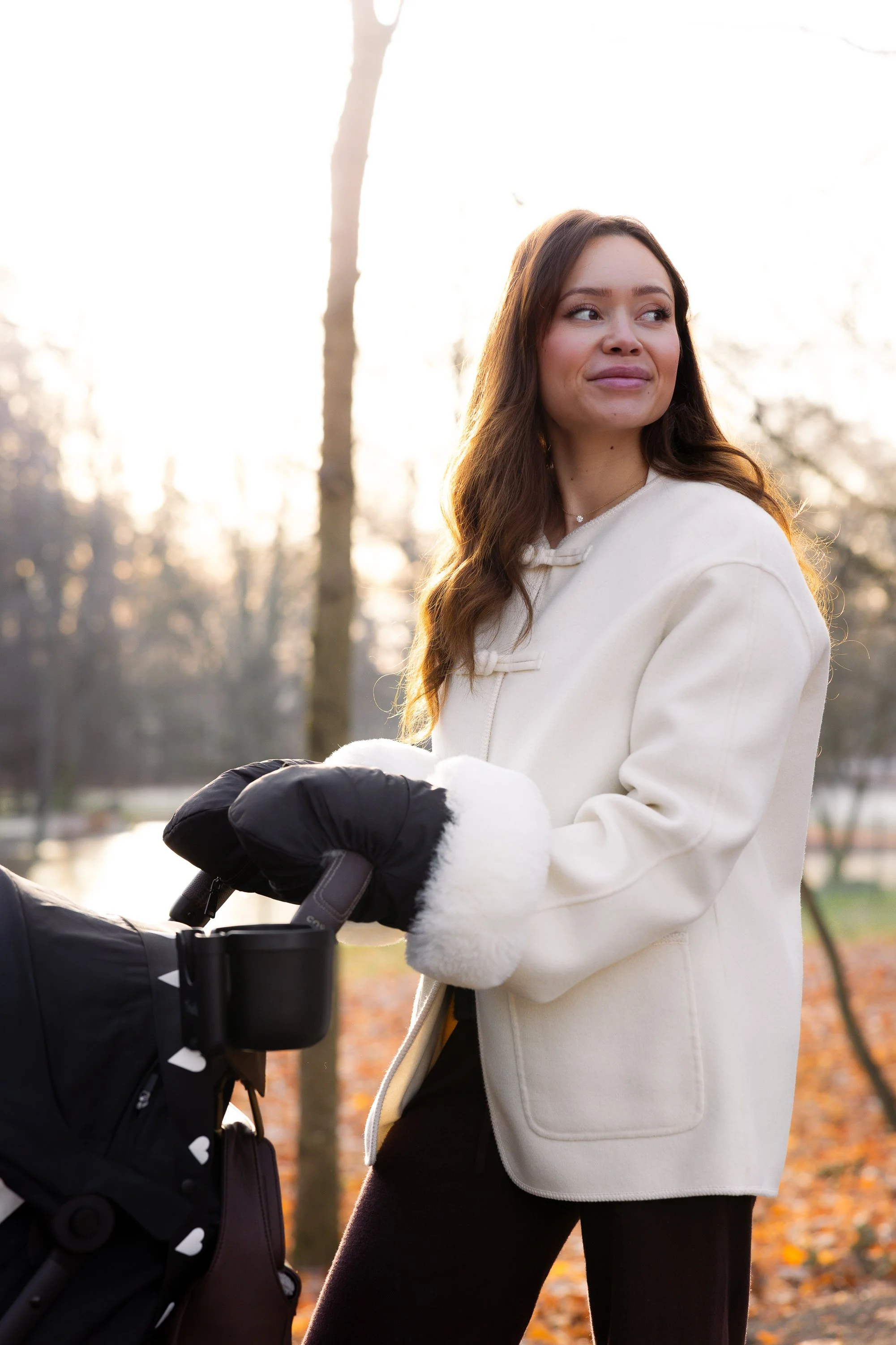 A woman in a white coat with black gloves standing outdoors with a stroller, trees in the background, during late afternoon or early evening.