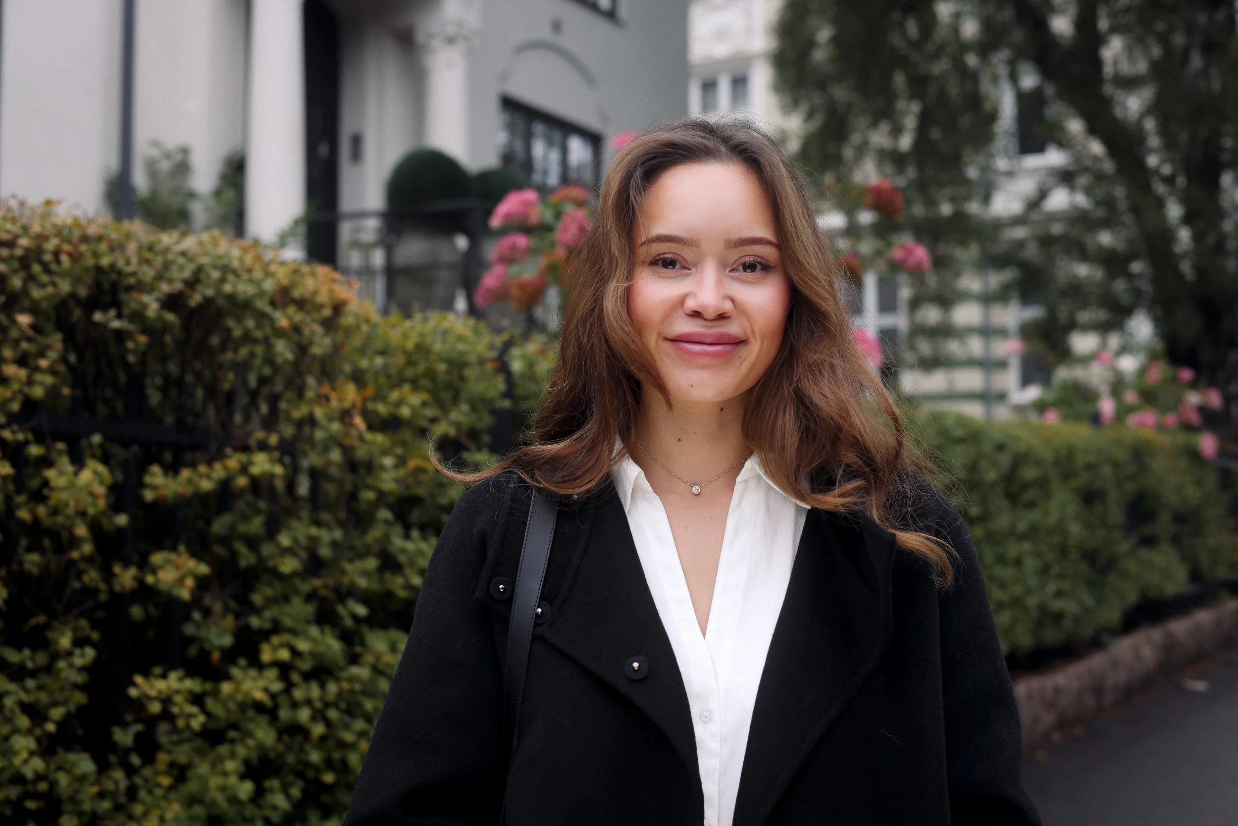 A young woman with wavy brown hair, wearing a white blouse and black coat, standing outdoors in front of a garden with bushes and pink flowers, smiling.