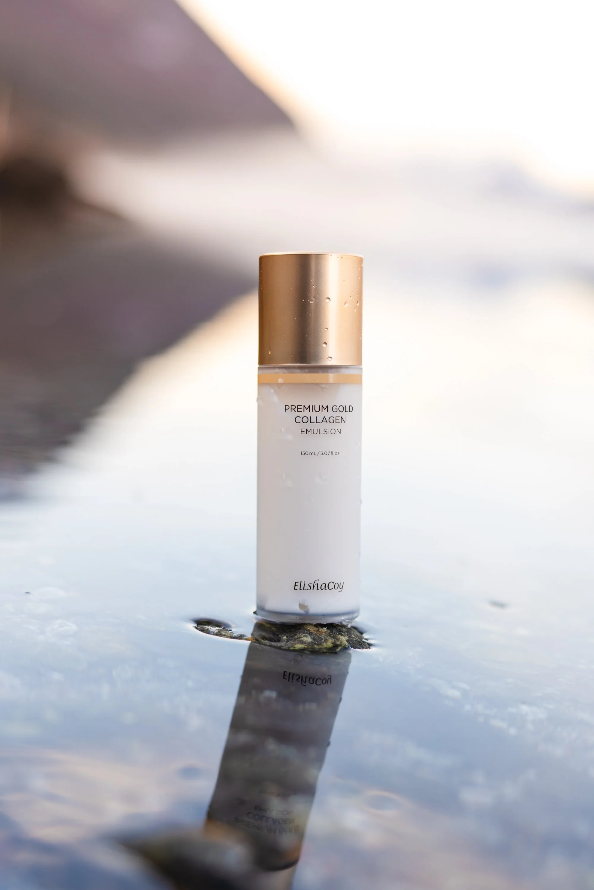 A bottle of ElishaCoy Premium Gold Collagen Emulsion standing on a wet rock, with water droplets on the bottle, reflected on the water surface, blurred natural background.