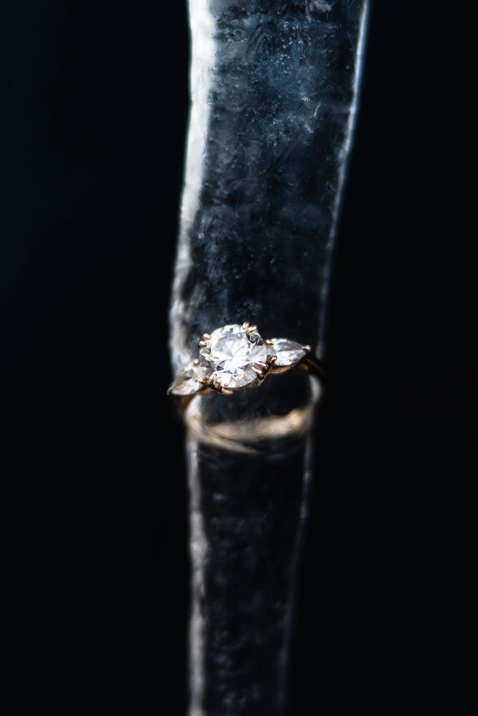 A gold ring with a large round diamond surrounded by small diamond accents, resting on a reflective black surface with a vertical icy or glass element behind it.