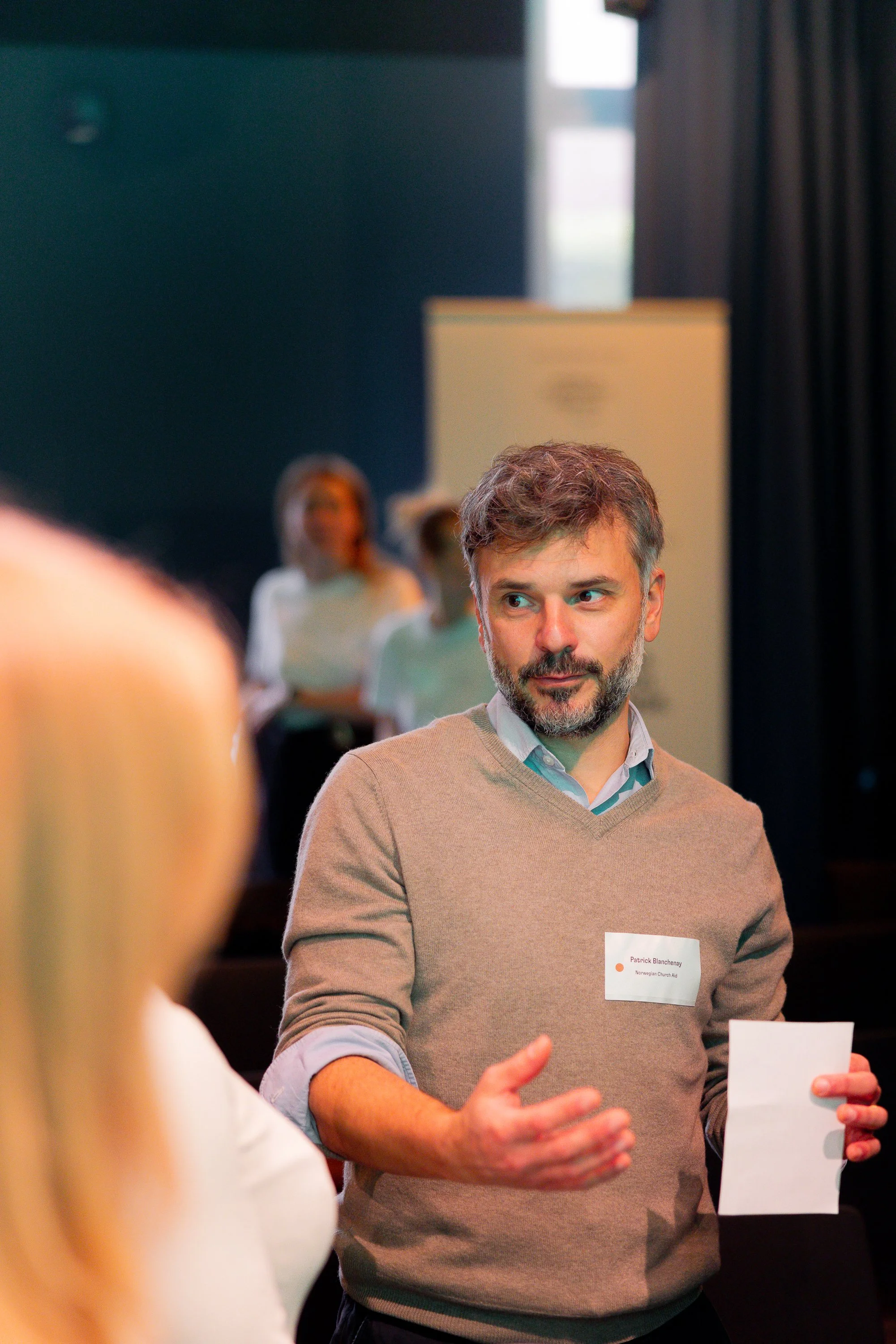A man with a beard and brown hair wearing a beige sweater over a light blue collared shirt, holding a white piece of paper in his left hand, appears to be gesturing with his right hand during a conversation in a conference or meeting setting. Blurred