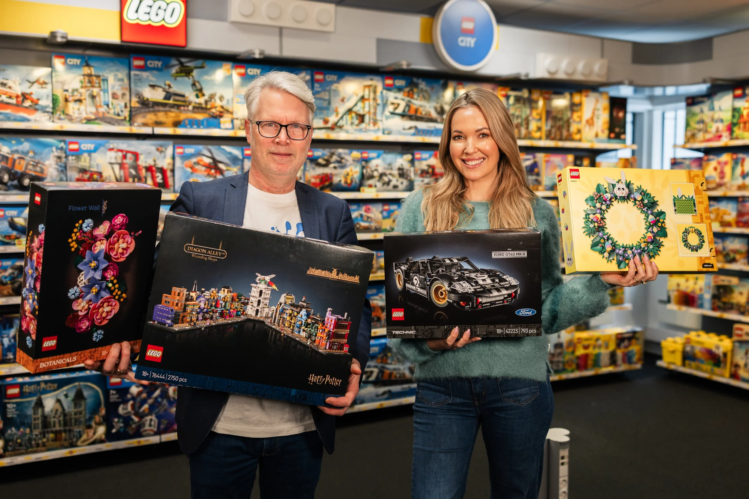 A man and woman shopping for LEGO sets in a toy store, holding multiple boxes including a floral-themed set, Harry Potter-themed set, racing car, and holiday wreath set.