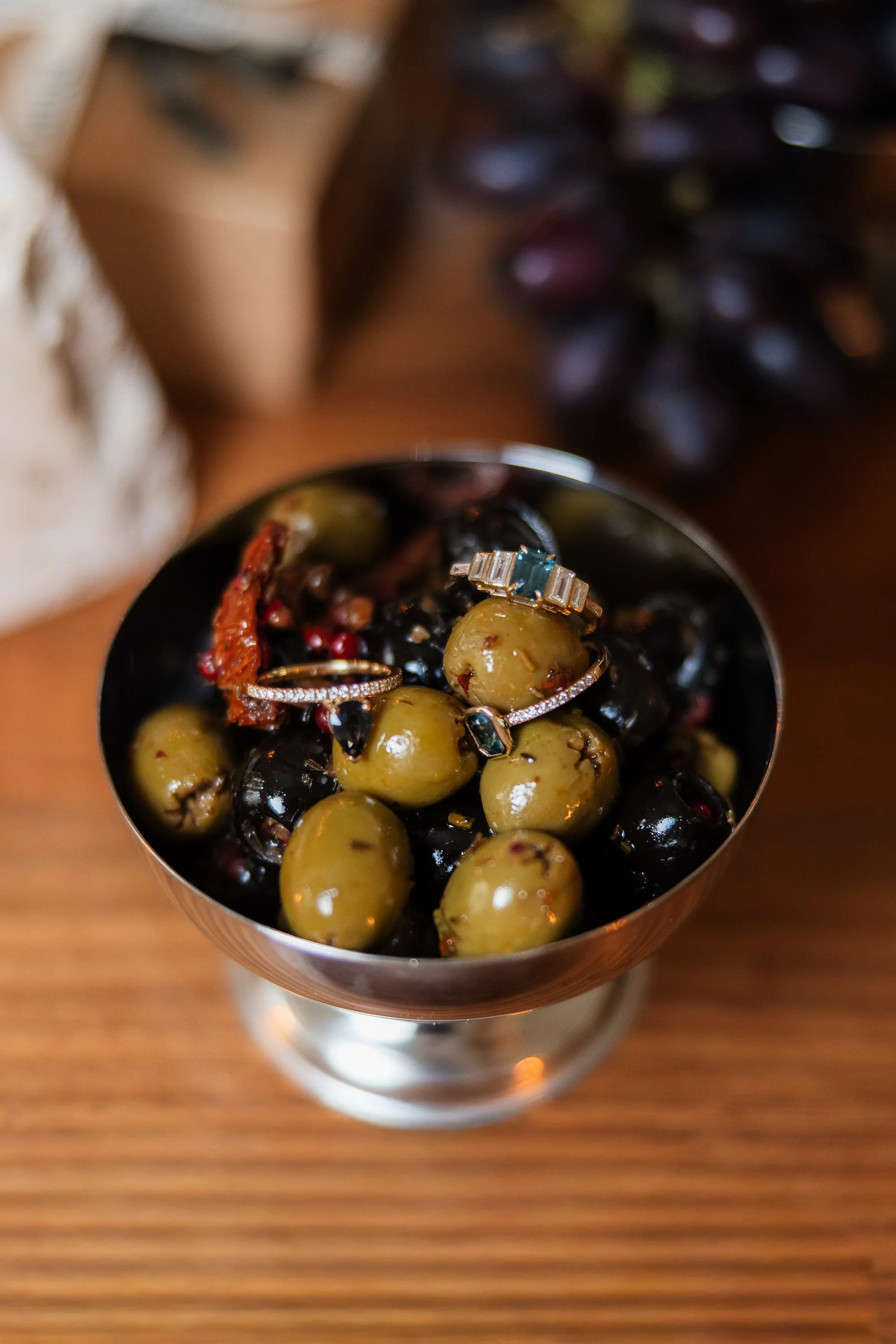 A metal bowl filled with green and black olives on a wooden surface. On top, there are jewelry rings, including a silver ring with a large blue stone, a gold ring with small clear stones, and a gold ring with a dark green stone.