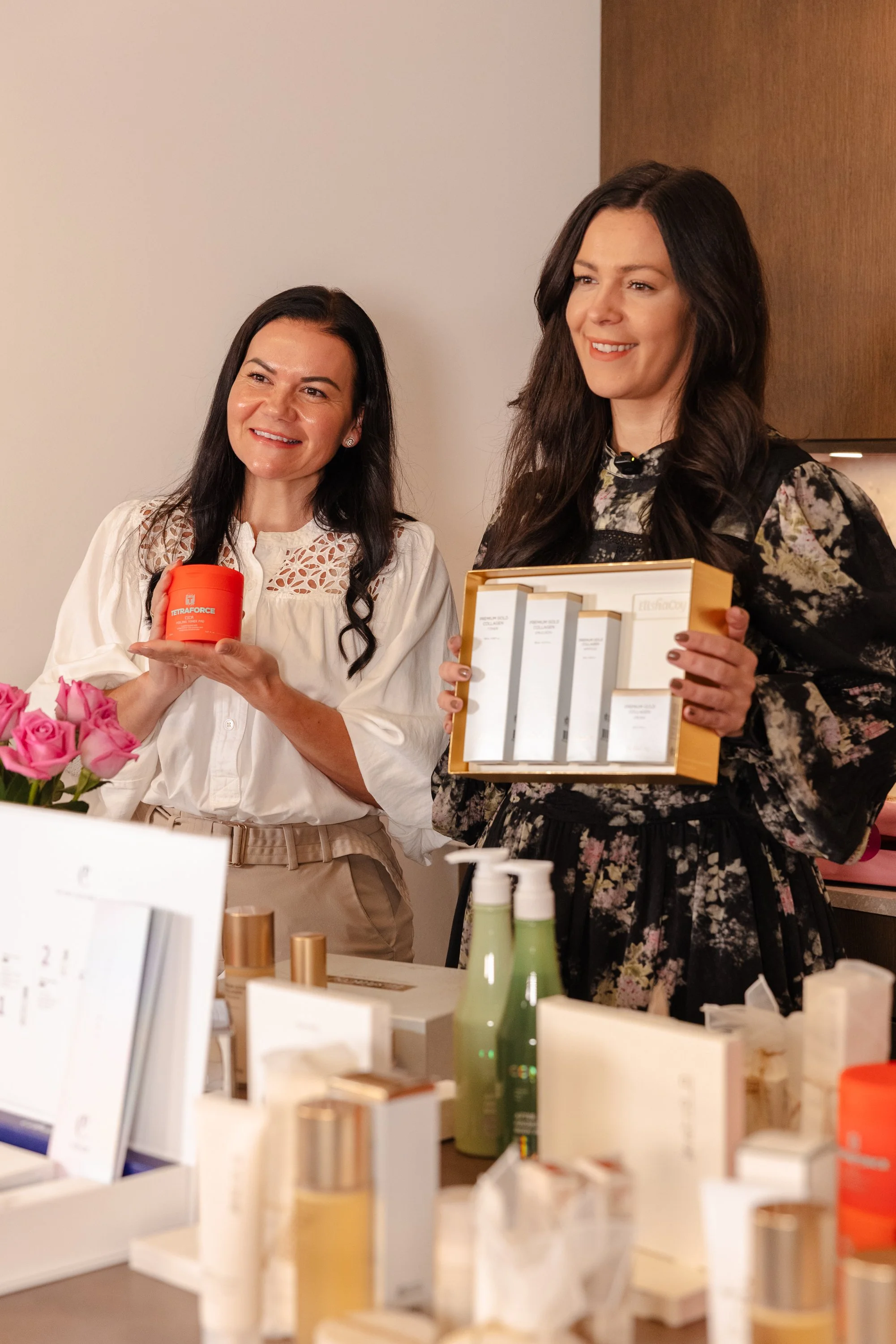 Two women standing indoors, smiling and holding skincare products at a display table with various beauty items and flowers.