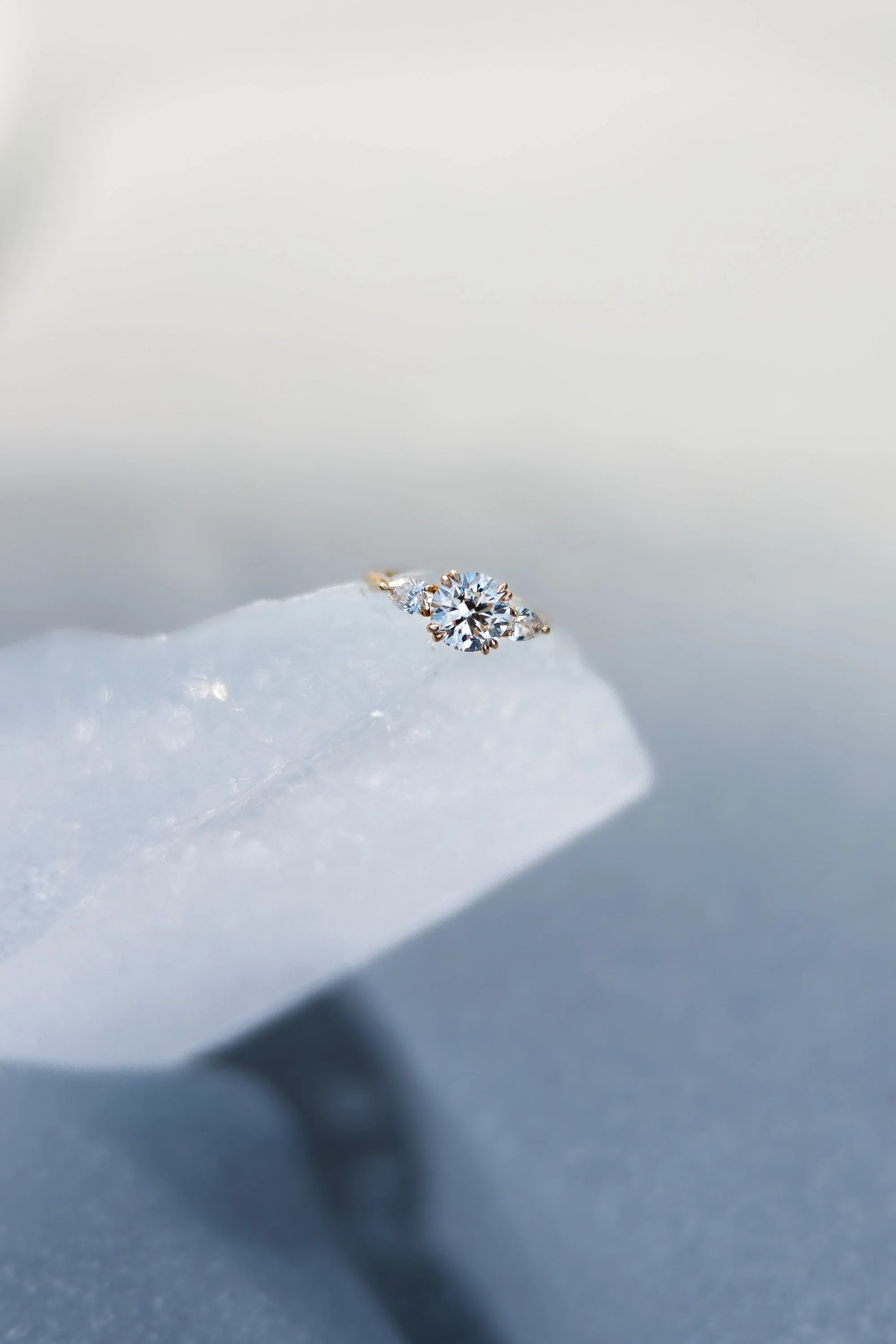 A diamond ring with a central round diamond flanked by two smaller diamonds, placed on a soft, textured white surface.