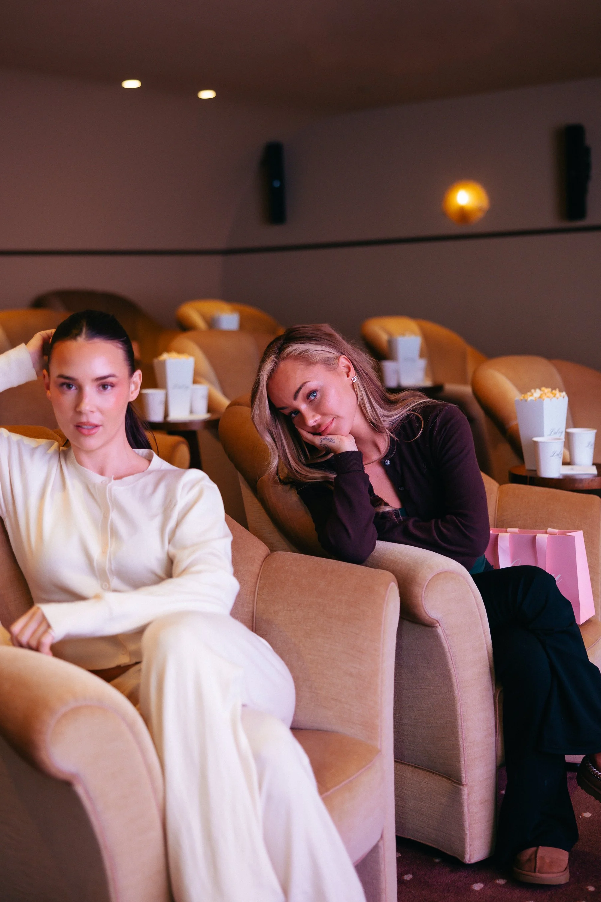 Two young women sitting in a movie theater, one wearing a white outfit and the other in black, with popcorn containers and pink shopping bag in the background.