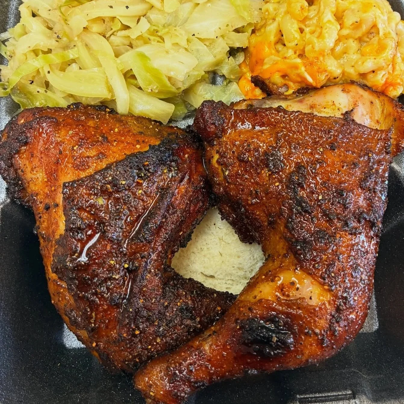 Two pieces of seasoned grilled chicken drumsticks, cooked with visible spices, served on a black tray with sides of cooked cabbage and creamy macaroni and cheese.