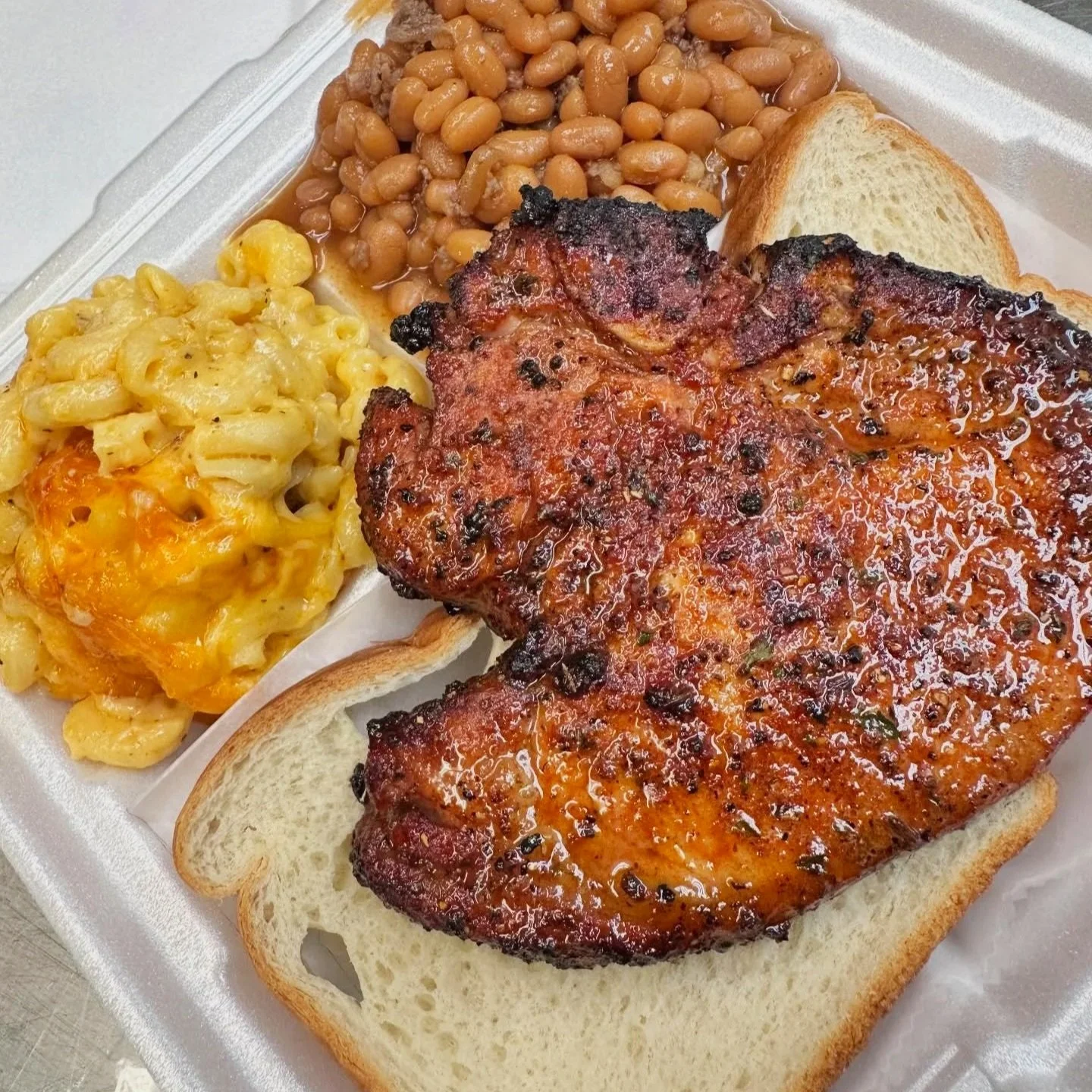 Barbecue pork chop with baked beans, macaroni and cheese, and a slice of white bread in a styrofoam takeout container.