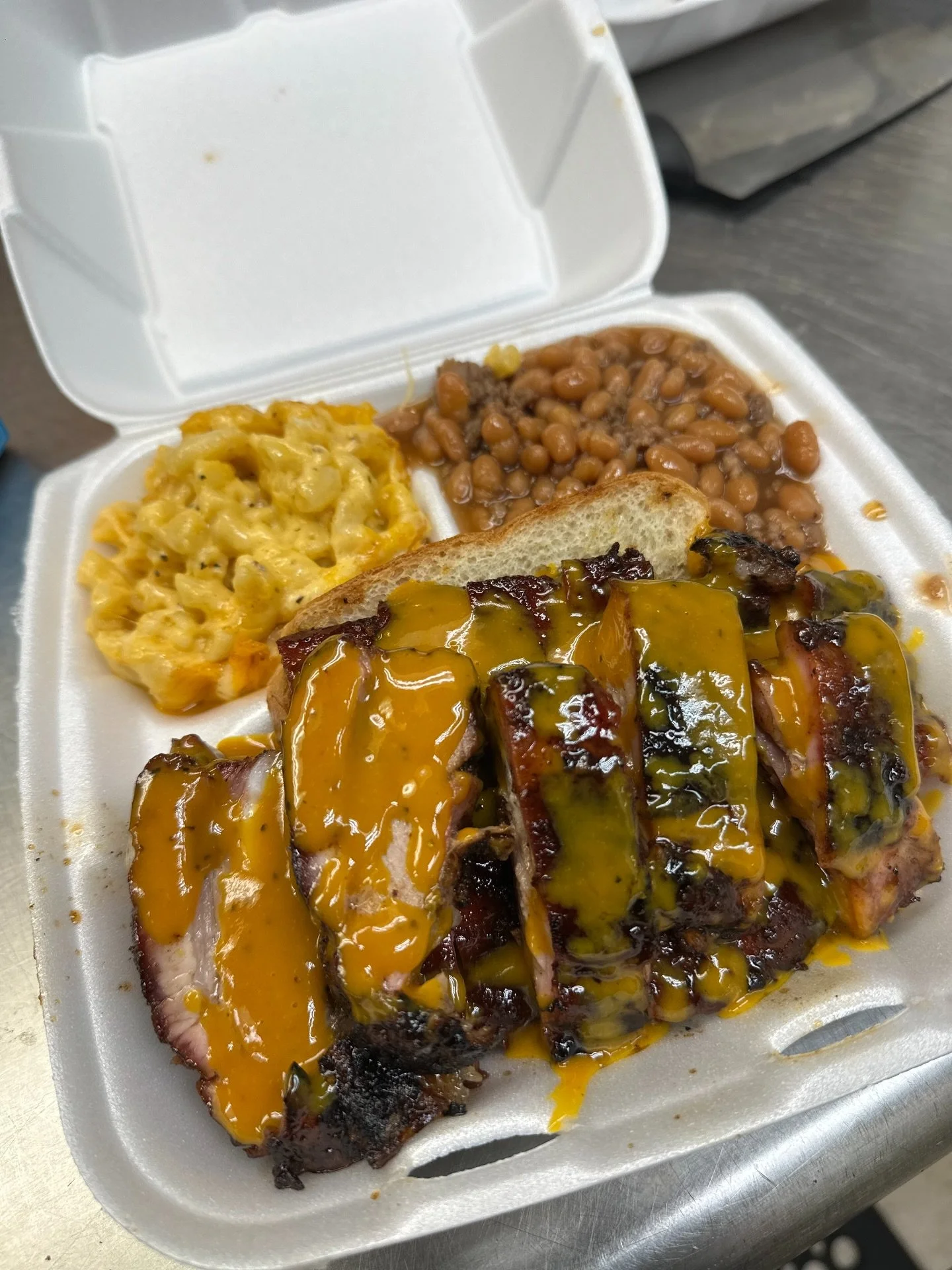 To-go styrofoam container with barbecue ribs topped with yellow sauce, macaroni and cheese, baked beans, and a slice of bread.