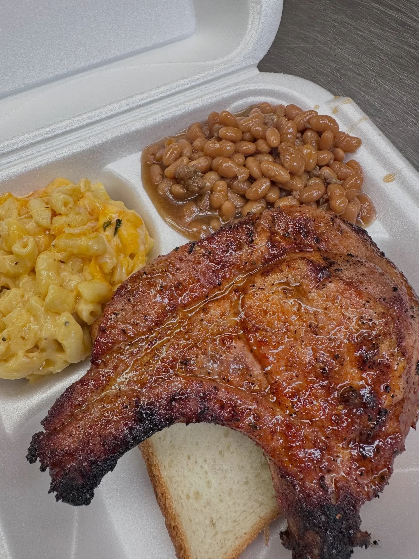 Barbecue pork chop with baked bread, baked beans, and macaroni and cheese on a styrofoam plate.