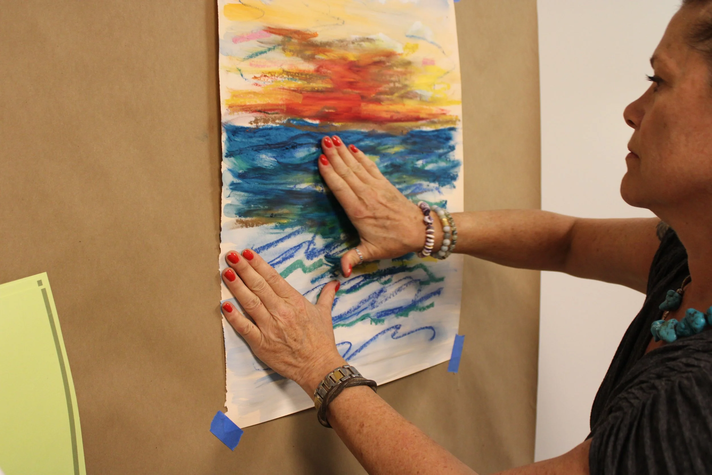 A woman with red polished nails, jewelry, and a black shirt is touching a colorful abstract painting with her hands. The painting features vibrant brushstrokes in yellow, red, blue, and green.