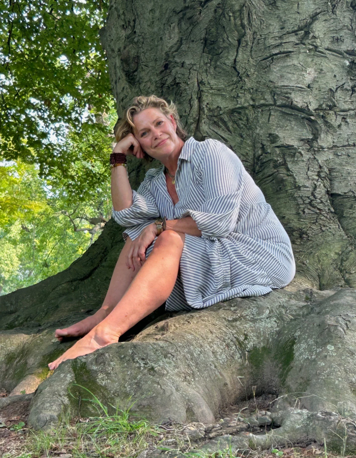 A woman sitting on a tree root, leaning against the trunk of a large tree in a lush green park or forest.