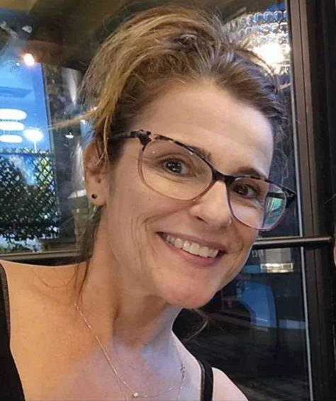 A woman with light brown hair and glasses smiling indoors near a window.