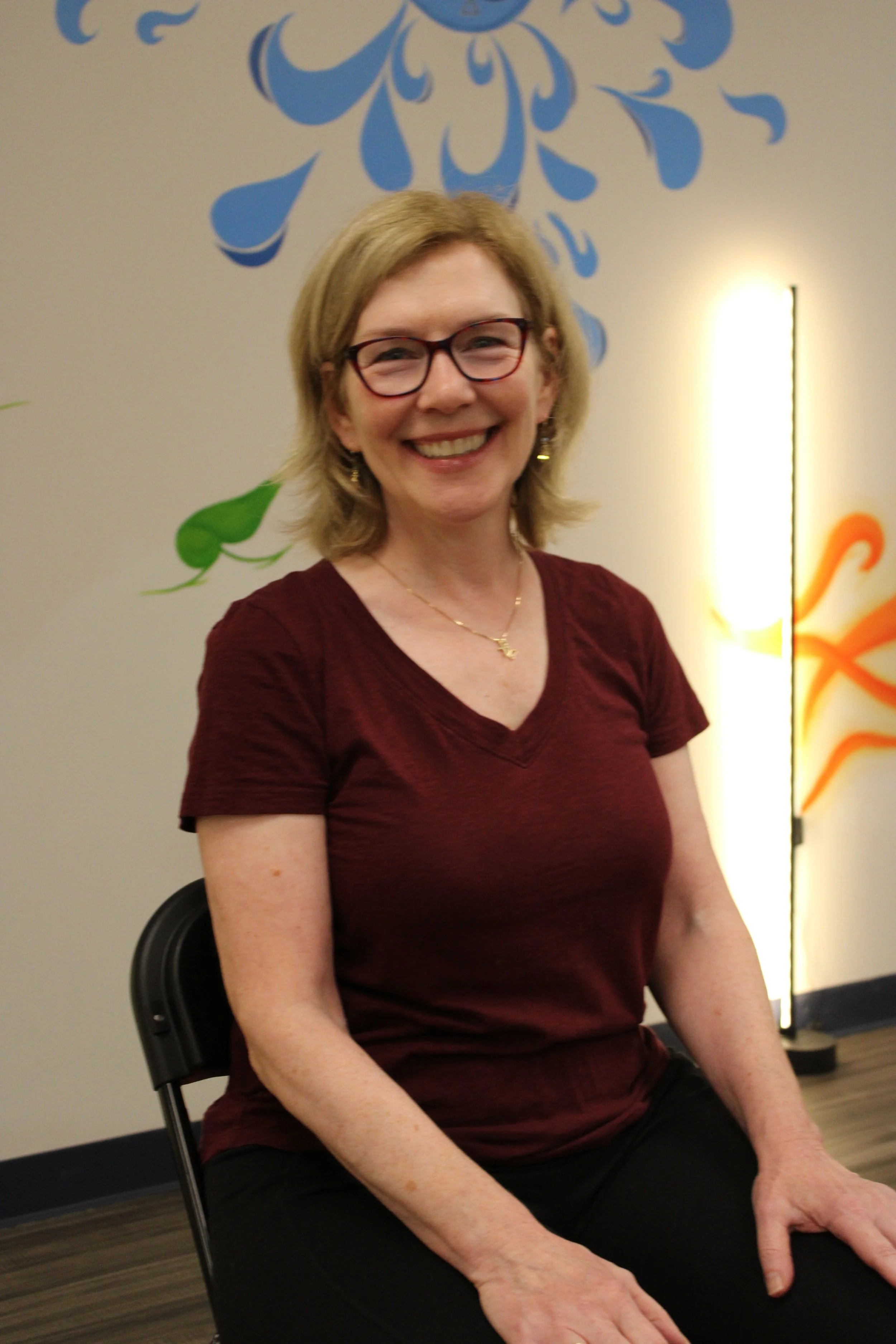 A woman with light brown hair, glasses, and a maroon shirt, smiling while sitting on a black chair in an indoor setting with a decorated wall and a light in the background.