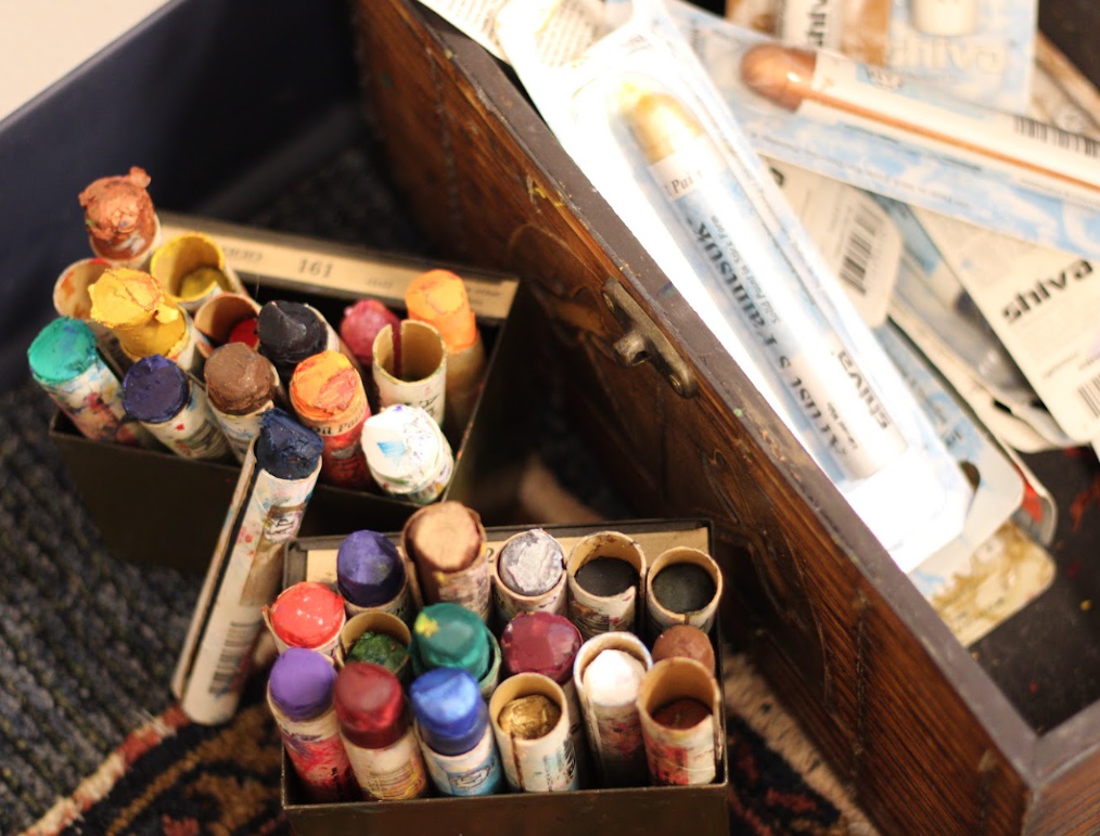 Two open tins filled with used pastel or chalk crayons, surrounded by blueprints and art supplies on a table.