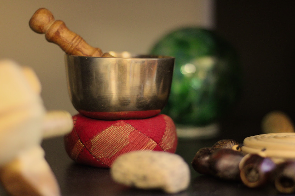 A metal bowl with a wooden pestle inside, placed on a red patterned fabric cushion, surrounded by seashells and with a blurred green object in the background.