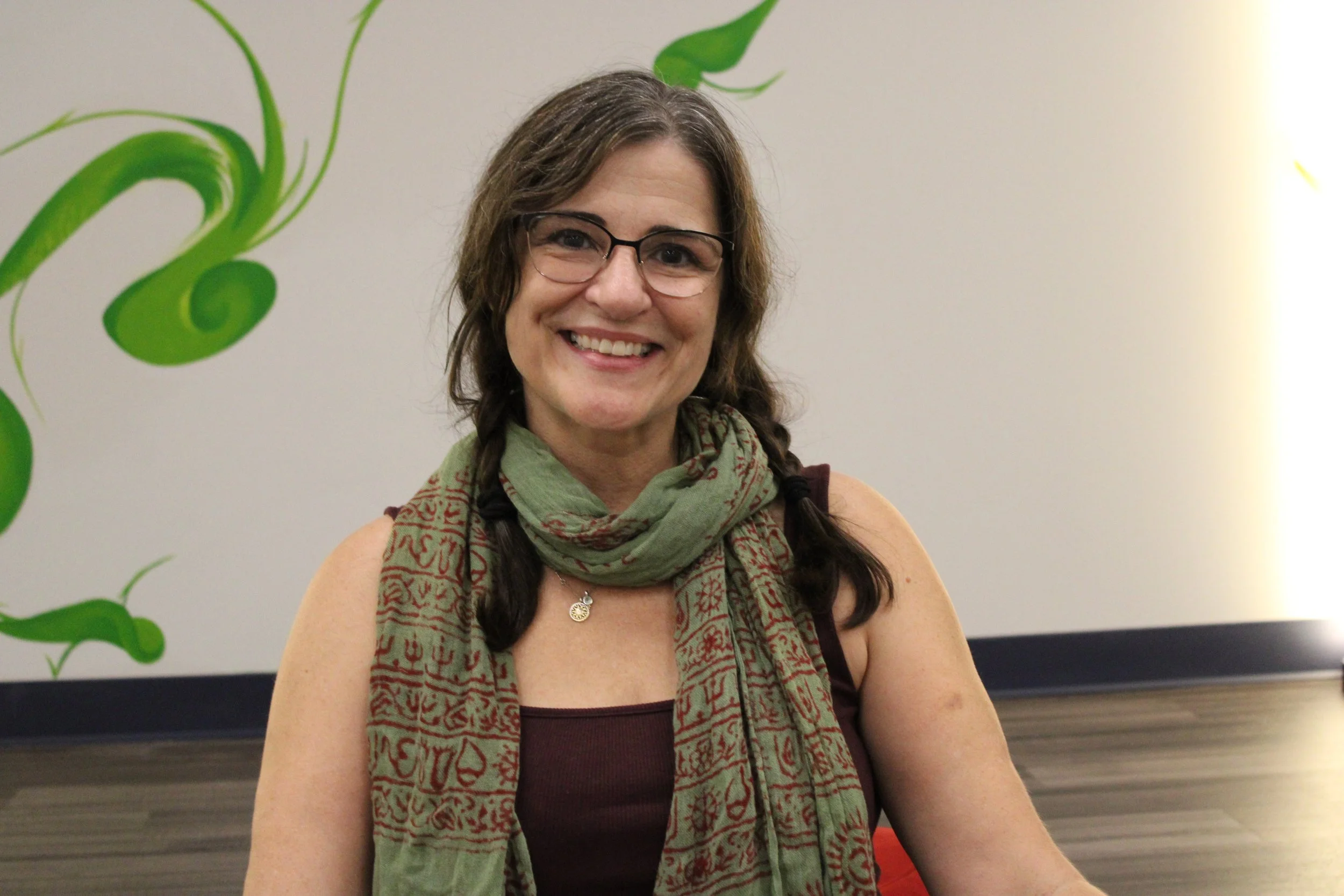 A smiling woman with glasses and long dark hair in braids, wearing a patterned scarf and tank top, sitting in front of a white wall with green abstract designs.
