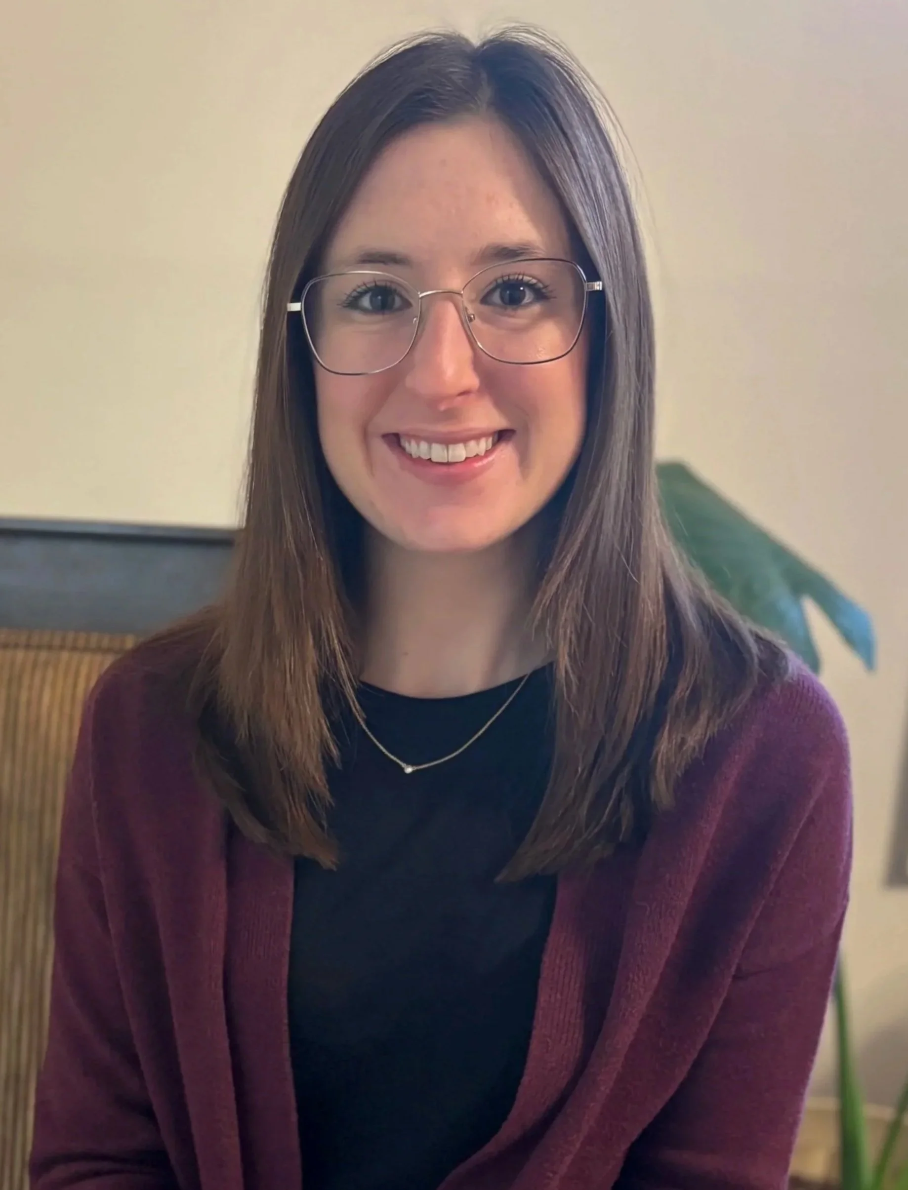 A smiling woman with shoulder-length brown hair, wearing glasses, a black top, a layered necklace, and a burgundy cardigan, sitting indoors with a plant and a piece of furniture in the background.