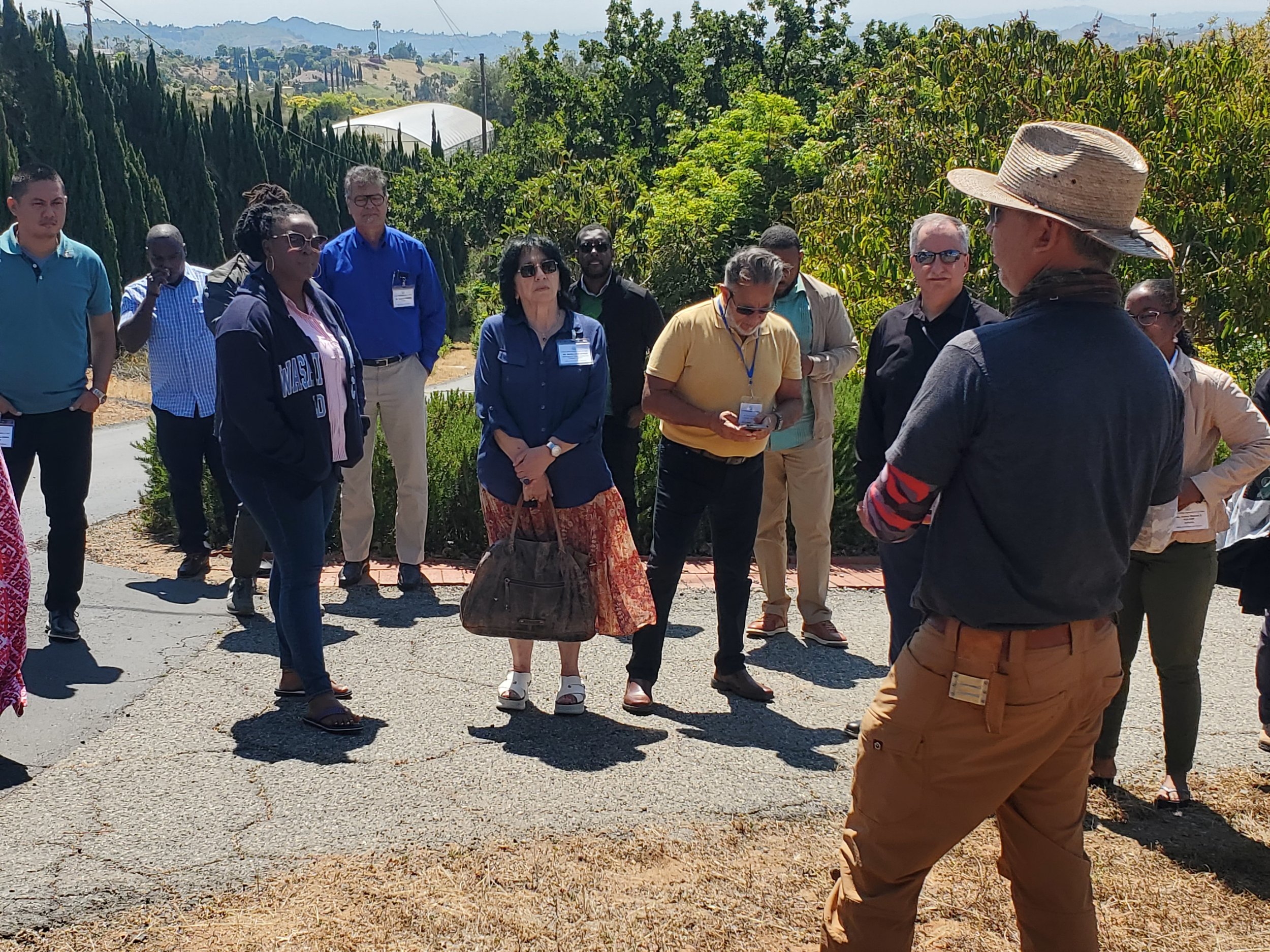 A group of people gathered outdoors on a sunny day, listening to a man wearing a straw hat and dark clothing, who is speaking to them. The background shows trees, bushes, and distant hills.