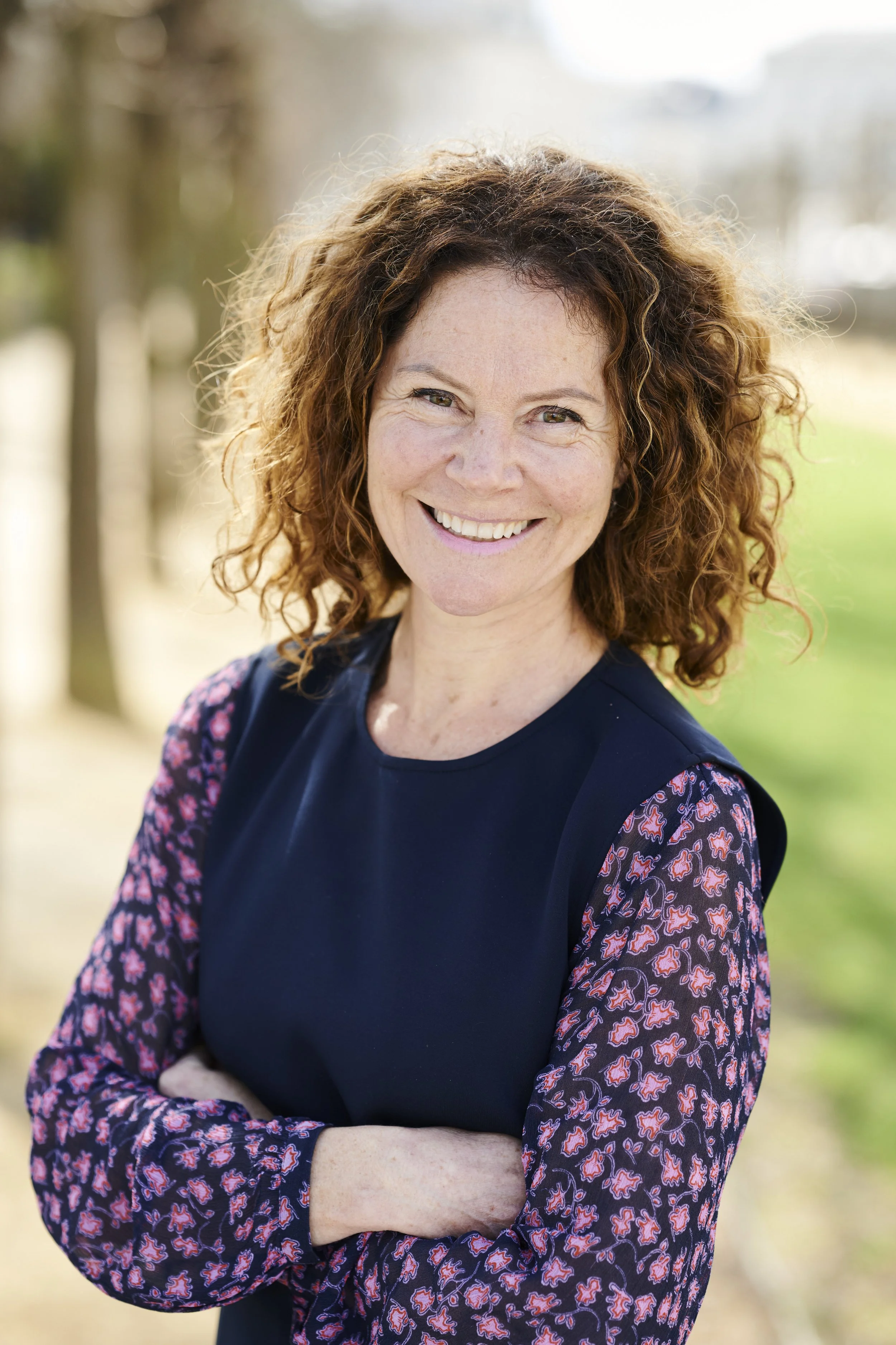 Headshot photo of a woman with curly red hair smiling outdoors, wearing a black top with floral patterned sleeves.