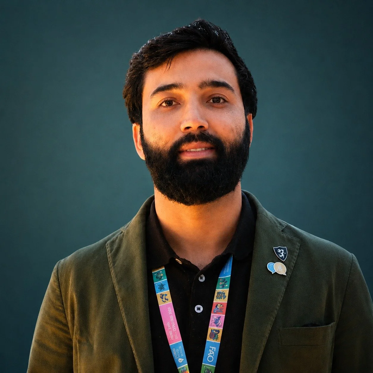 Headshot photo of a man with dark hair and a beard wearing a jacket and a black shirt, with a lanyard around his neck, standing against a teal background.