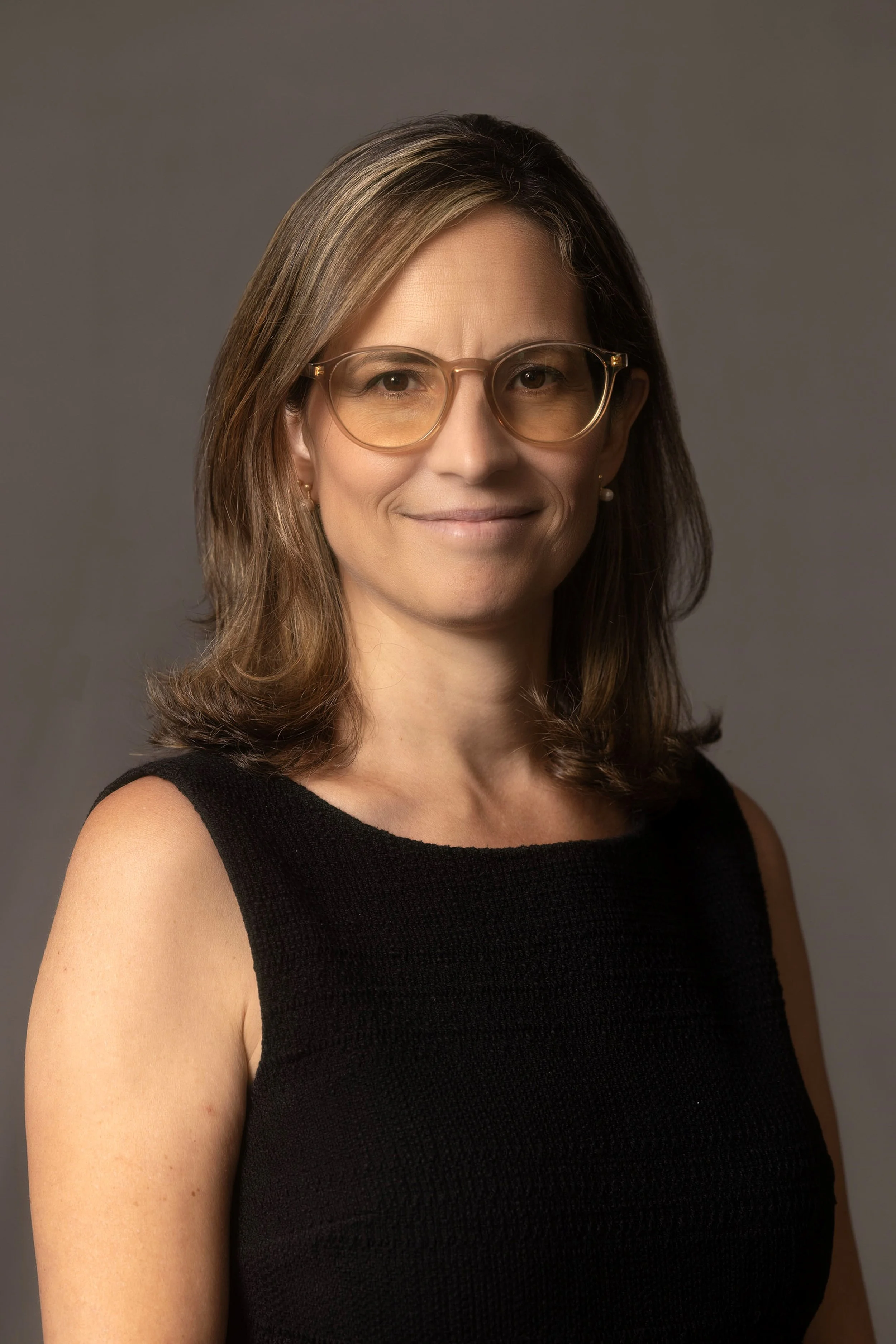 Headshot photo of a woman with shoulder-length brown hair and glasses, wearing a black top, smiling against a gray background.