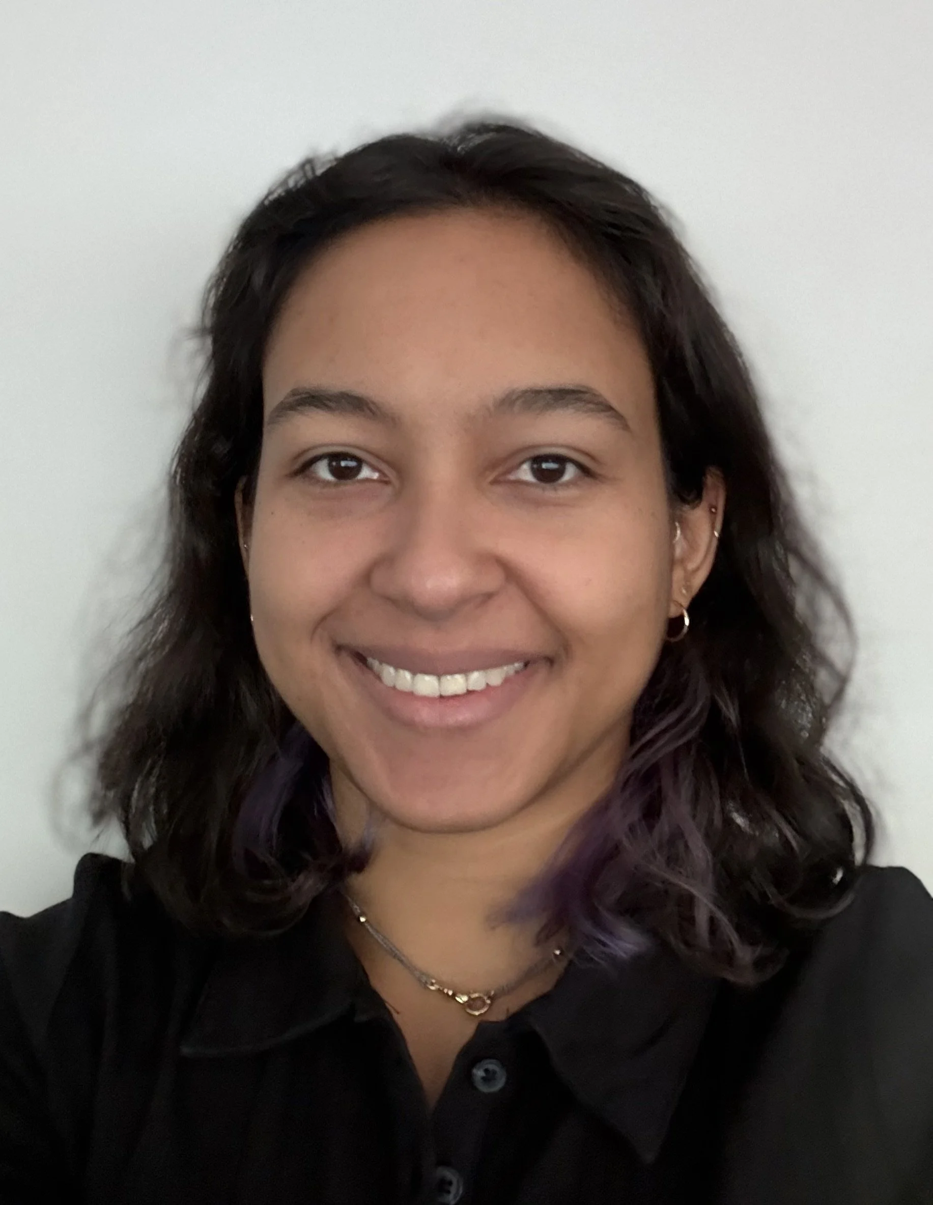 Headshot photo of a woman with dark wavy hair and purple highlights, wearing earrings and a black button-up shirt, against a plain white background.