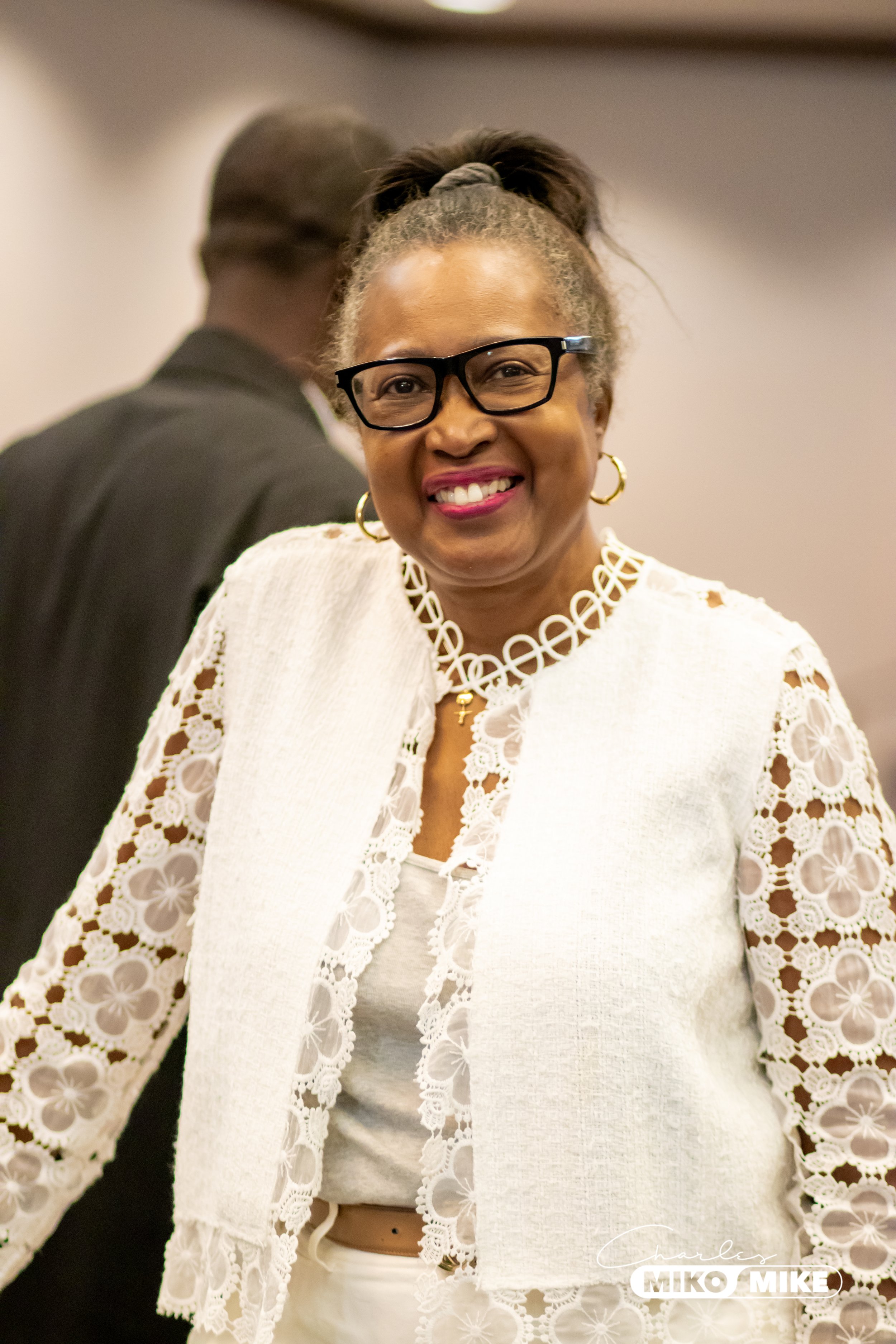 Headshot photo of a woman with glasses and earrings wearing a white lace outfit at an indoor event.