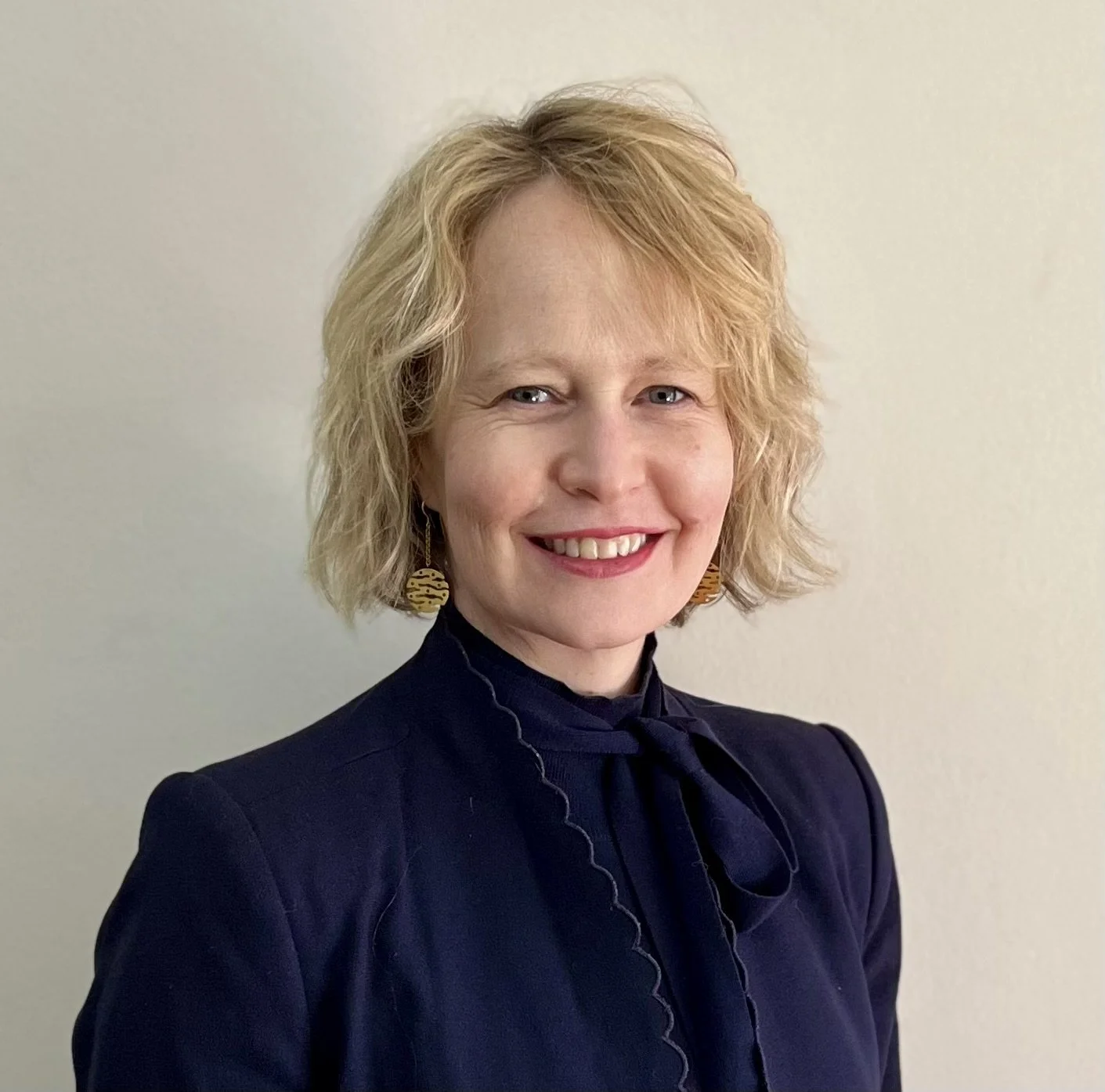 Headshot photo of a woman with short blonde hair, wearing a dark blazer and gold earrings, standing against a plain light background.