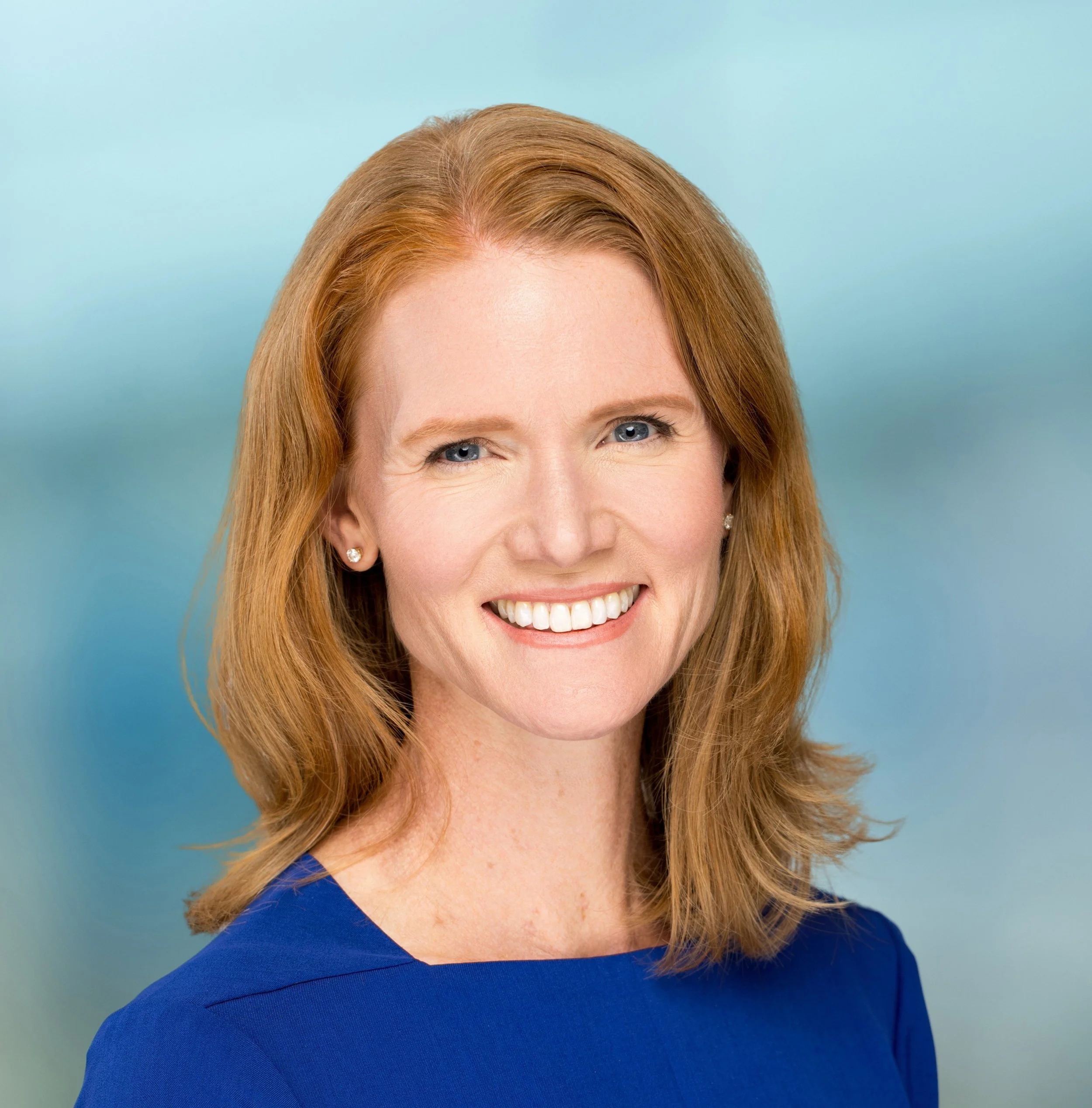 Headshot of a woman with blue eyes, red hair, and wearing a blue top, against a light blue background.