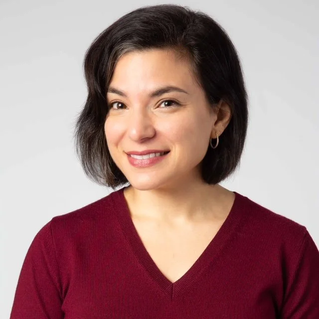 Headshot photo of a woman with shoulder-length dark hair, wearing a maroon V-neck sweater and small hoop earrings, against a plain light gray background.