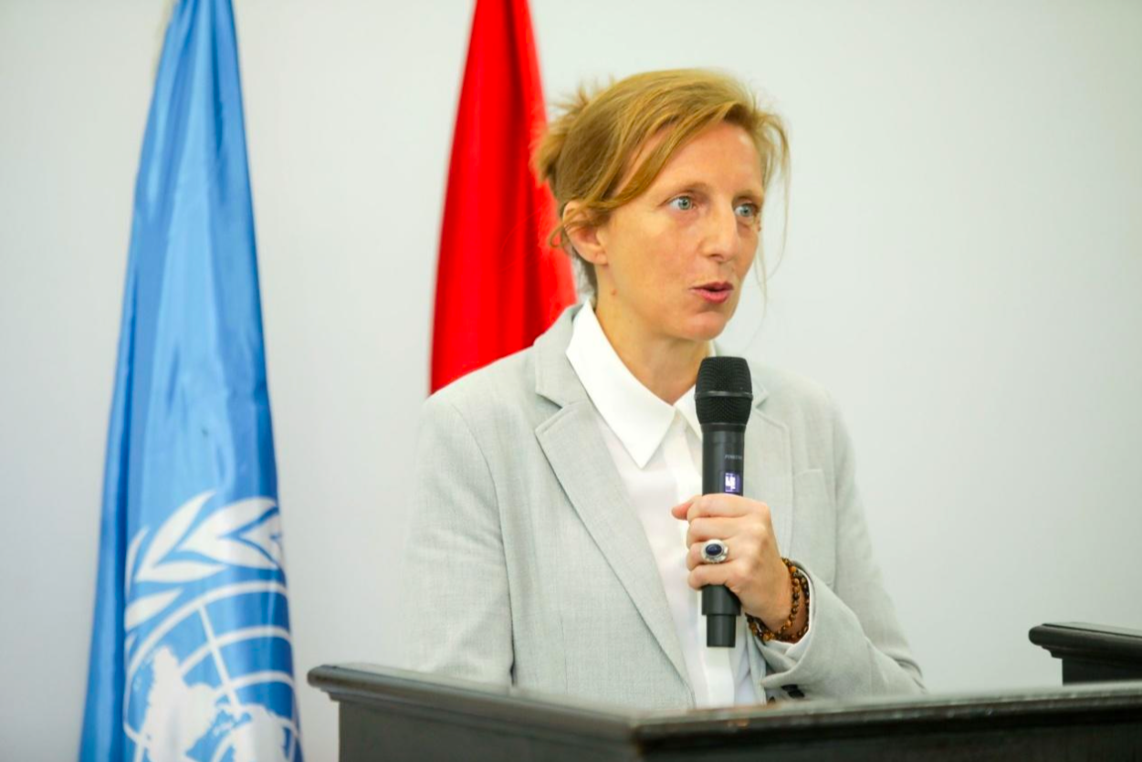 A woman with short blonde hair in a light gray blazer and white shirt speaking into a black microphone at a podium, with a UN flag and a red flag behind her.