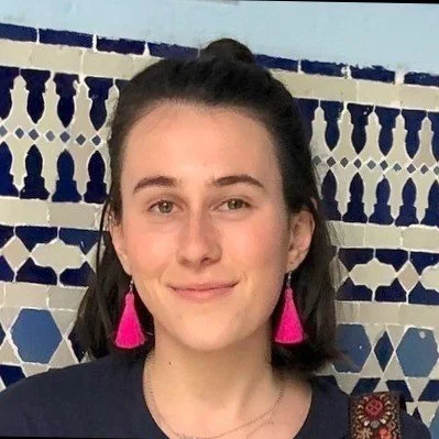 Headshot photo of a woman with dark hair wearing pink tassel earrings and a navy top standing in front of a decorative blue and white tiled wall.