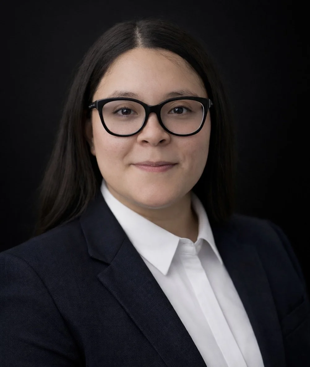 Headshot photo of a woman with dark hair, wearing glasses, a white shirt, and a dark blazer, posing against a black background.