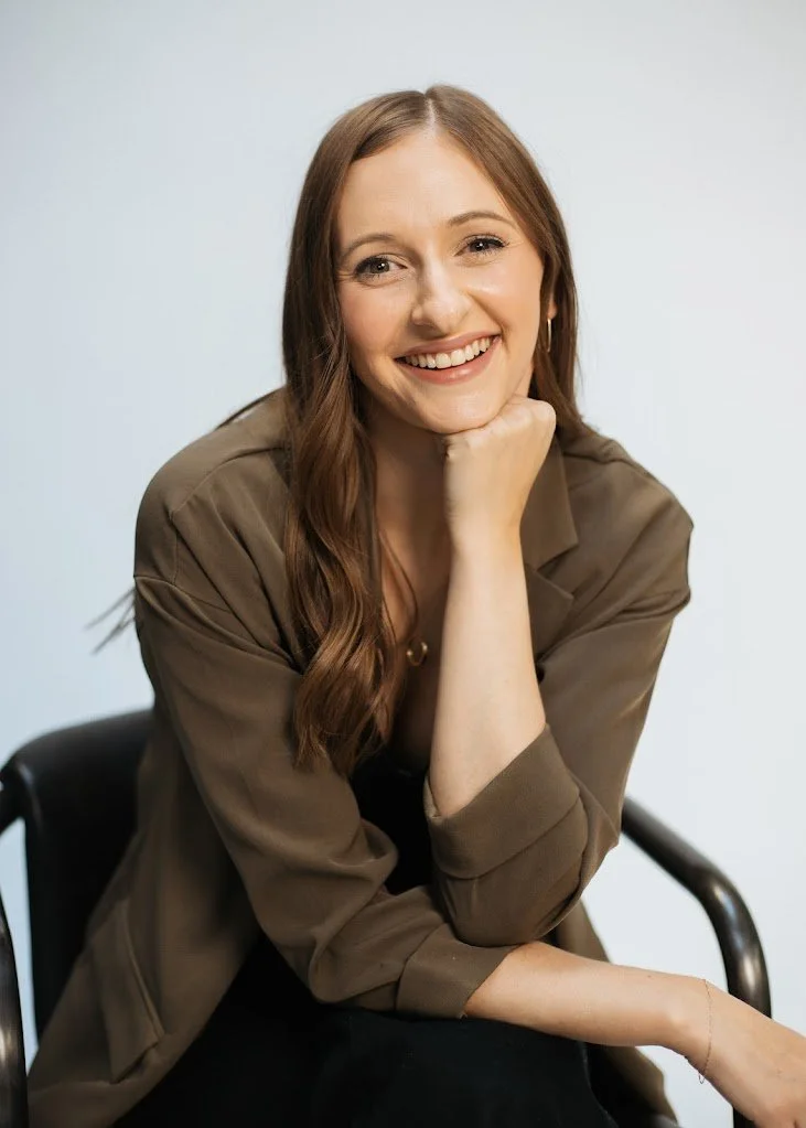 Shannon O'Keefe, Registered Clinical Counsellor (RCC), smiling warmly while resting her chin on her hand. She is wearing a green blazer and sitting in a black chair against a light background.