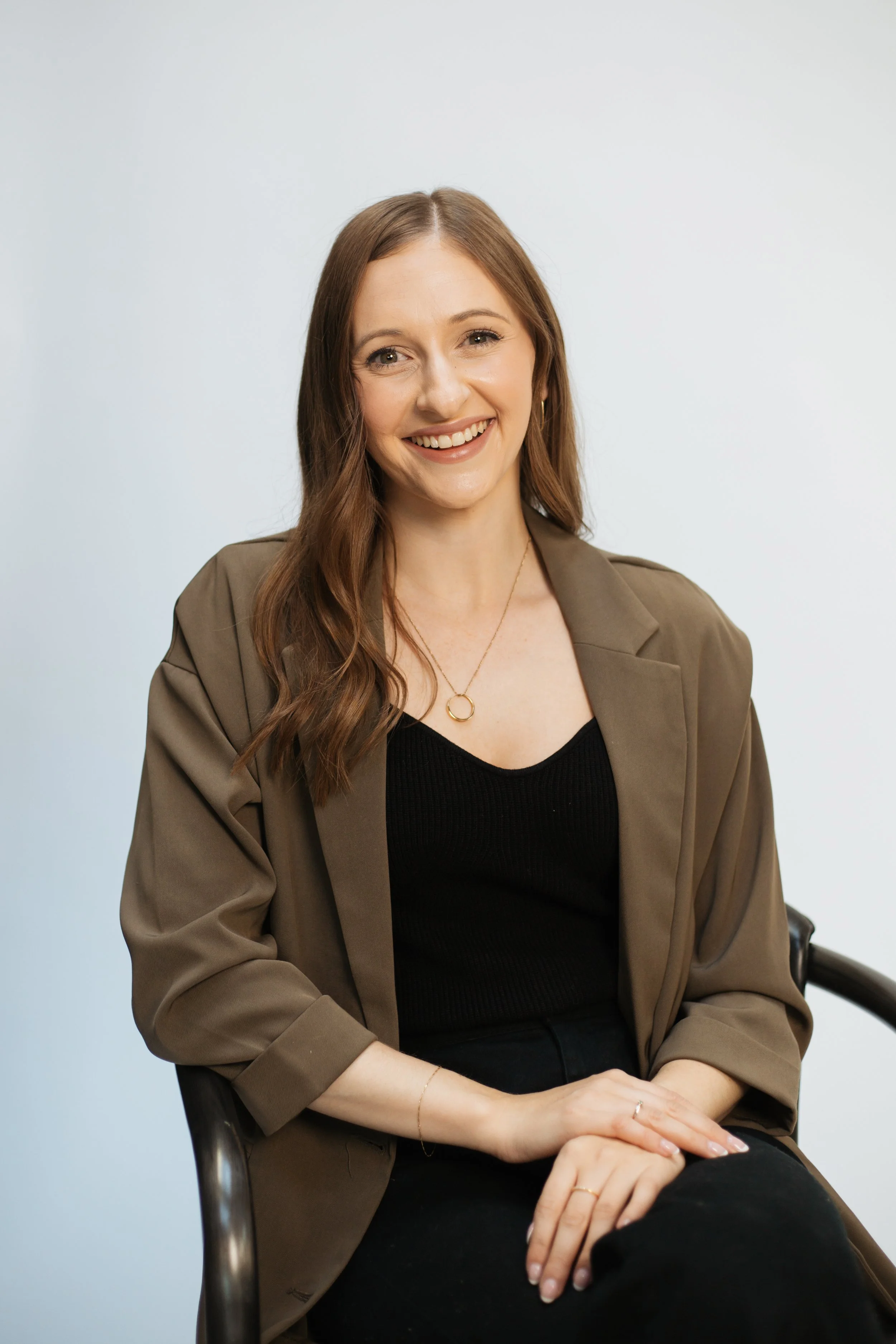 Shannon O'Keefe, RCC, sitting on a metal chair and smiling warmly at the camera. She has long wavy brown hair and wears a professional brown blazer over a black top against a light background.