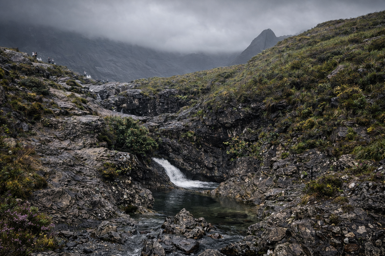 A rugged mountainous landscape with dark rocks, a small waterfall, and a stream in the foreground. Green bushes and plants grow among the rocks, and misty clouds hover over the peaks in the background.
