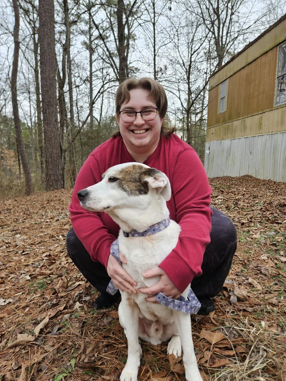 A owner and operator of this Chattanooga pet sitting company, crouching outdoors in a wooded area, holding a large white and brown dog with a purple collar and Bandana.