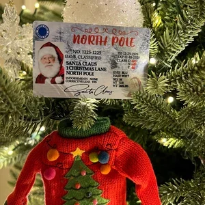 A Christmas tree ornament depicting a Santa ticket and a red sweater with a Christmas tree design on it hanging on a Christmas tree branch.