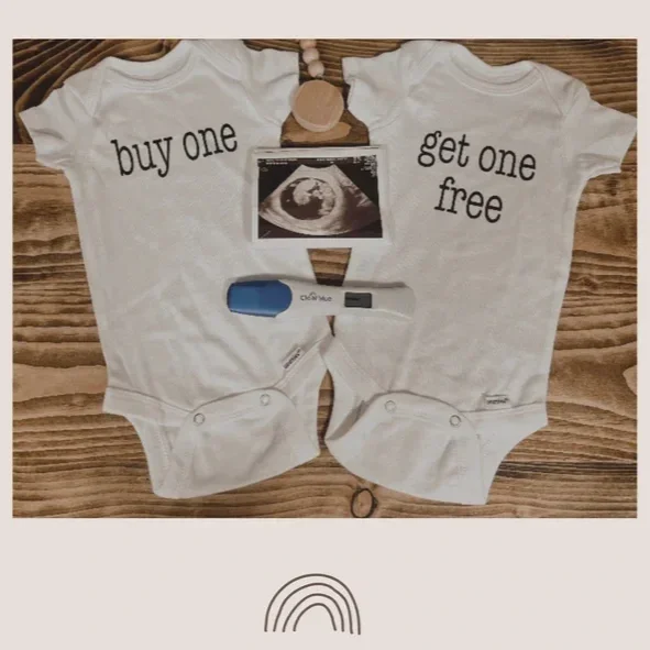 A baby announcement display with two white baby bodysuits. The left bodysuit says 'buy one' and the right says 'get one free.' There is an ultrasound picture of a baby, a pregnancy test, and a pill bottle placed above the bodysuits.