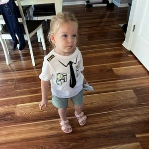 Young girl standing on a wooden floor in a home, wearing a white shirt with black tie design, shorts, and sandals.