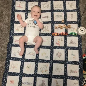 Baby lying on a quilt with embroidered squares, surrounded by small toy cars and a few toy blocks.