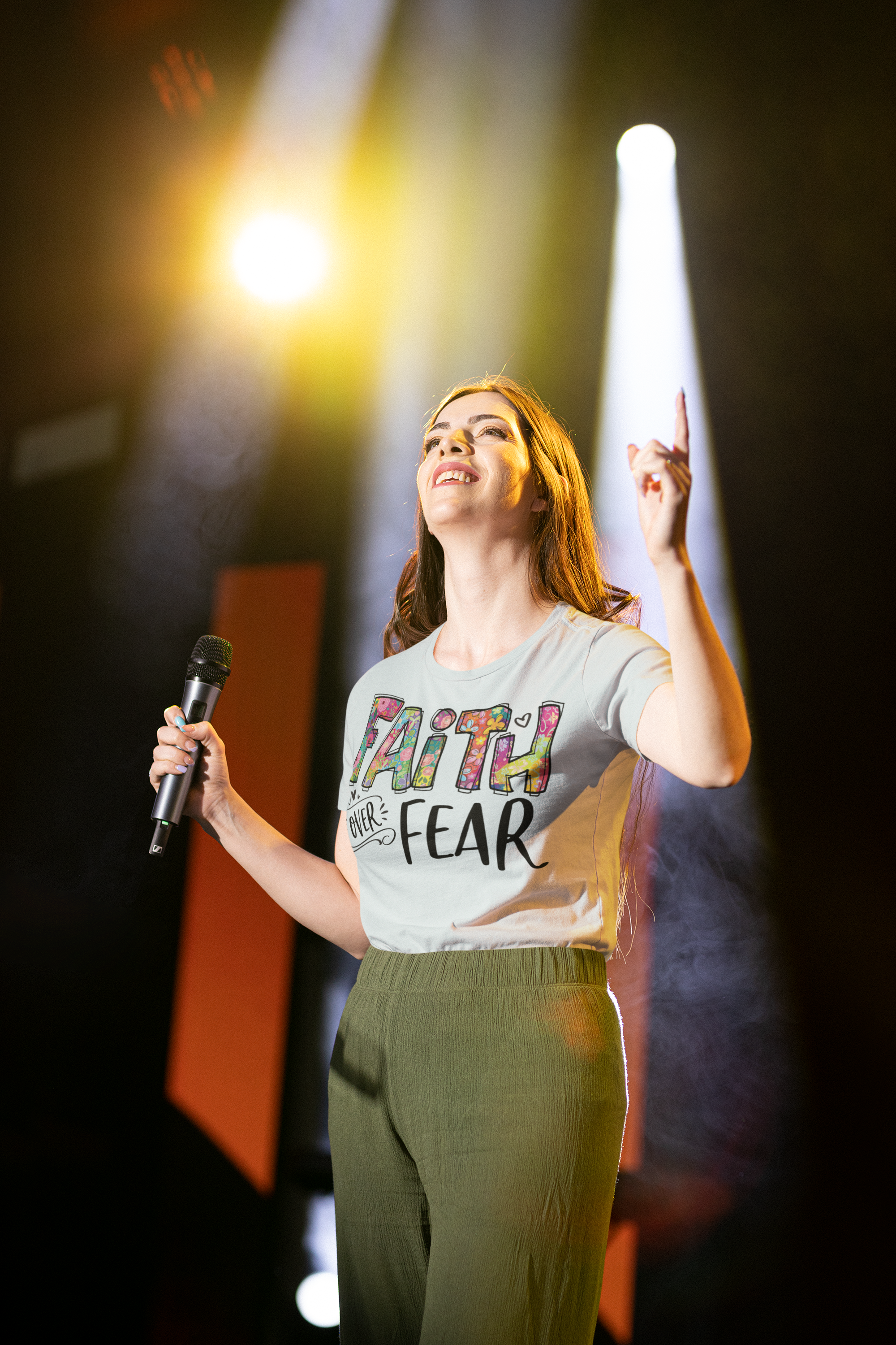A woman on stage holding a microphone in her right hand, smiling, and gesturing upward with her left hand. She wears a graphic T-shirt with colorful text that says 'FAITH OVER FEAR' and green pants. Bright stage lights illuminate her from behind.