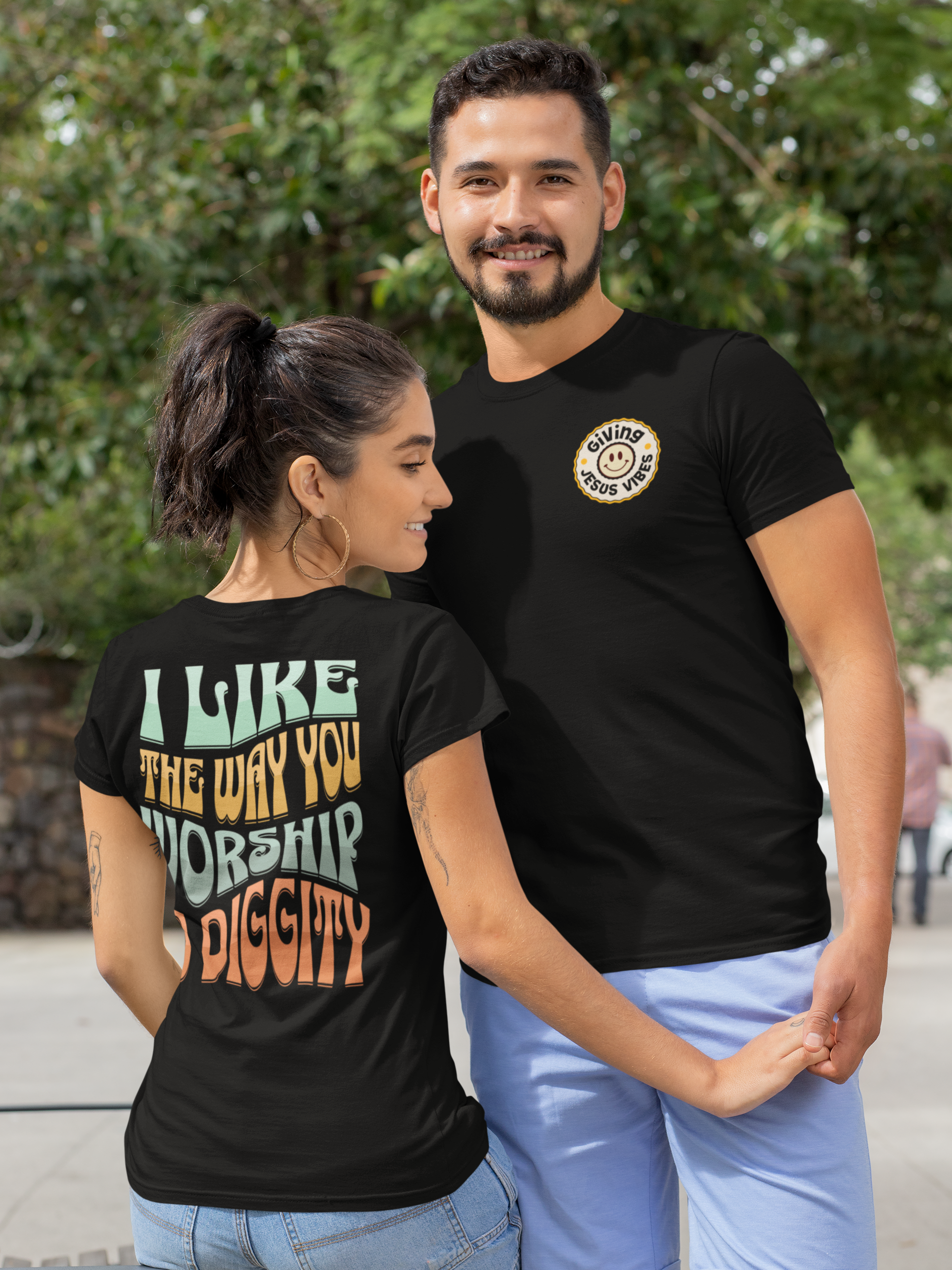 A smiling young woman with a ponytail and hoop earrings standing next to a tall young man with a beard, both wearing black T-shirts with positive messages, outdoors with green trees in the background.