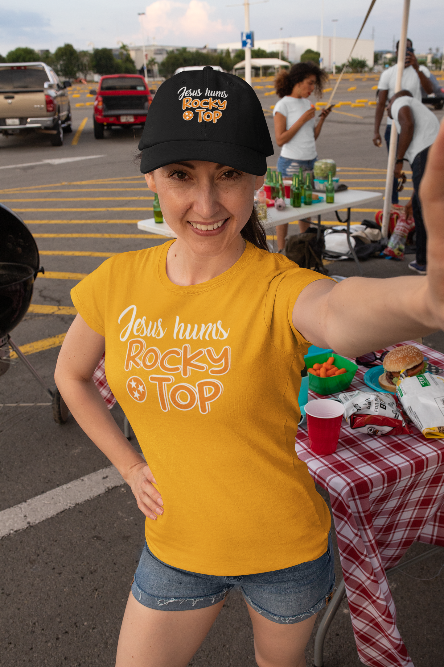 A woman smiling and taking a selfie at an outdoor gathering in a parking lot. She is wearing a black cap and a yellow t-shirt with the text 'Jesus hums Rocky Top'. Behind her are people, tables with snacks, and parked cars.