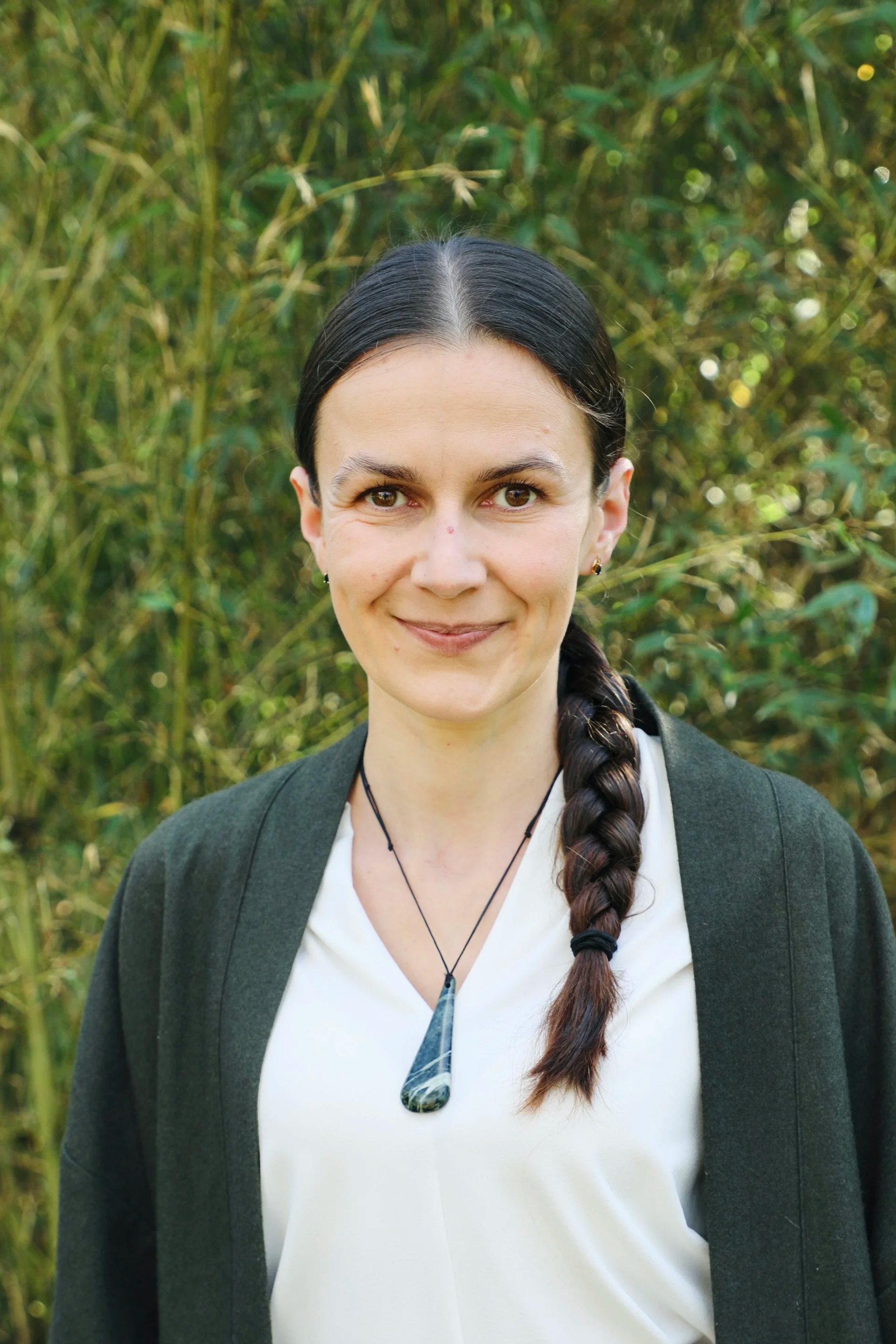 A woman with long dark braided hair, wearing a dark blazer and a white top, standing outdoors with green foliage in the background.