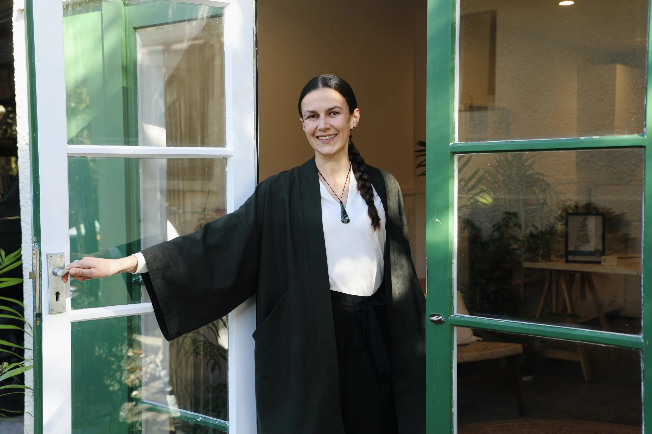 A woman with dark hair in a braid smiling and opening a green door with glass panels, standing inside a room with plants and a framed picture inside.