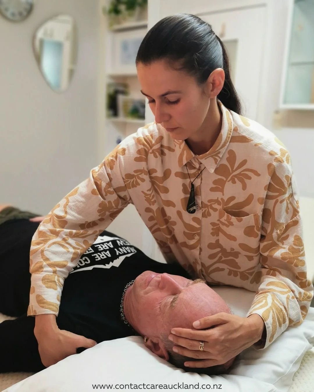 A woman providing first aid or medical assistance to a man lying on a bed, with her hand on his forehead, in a clinical setting.