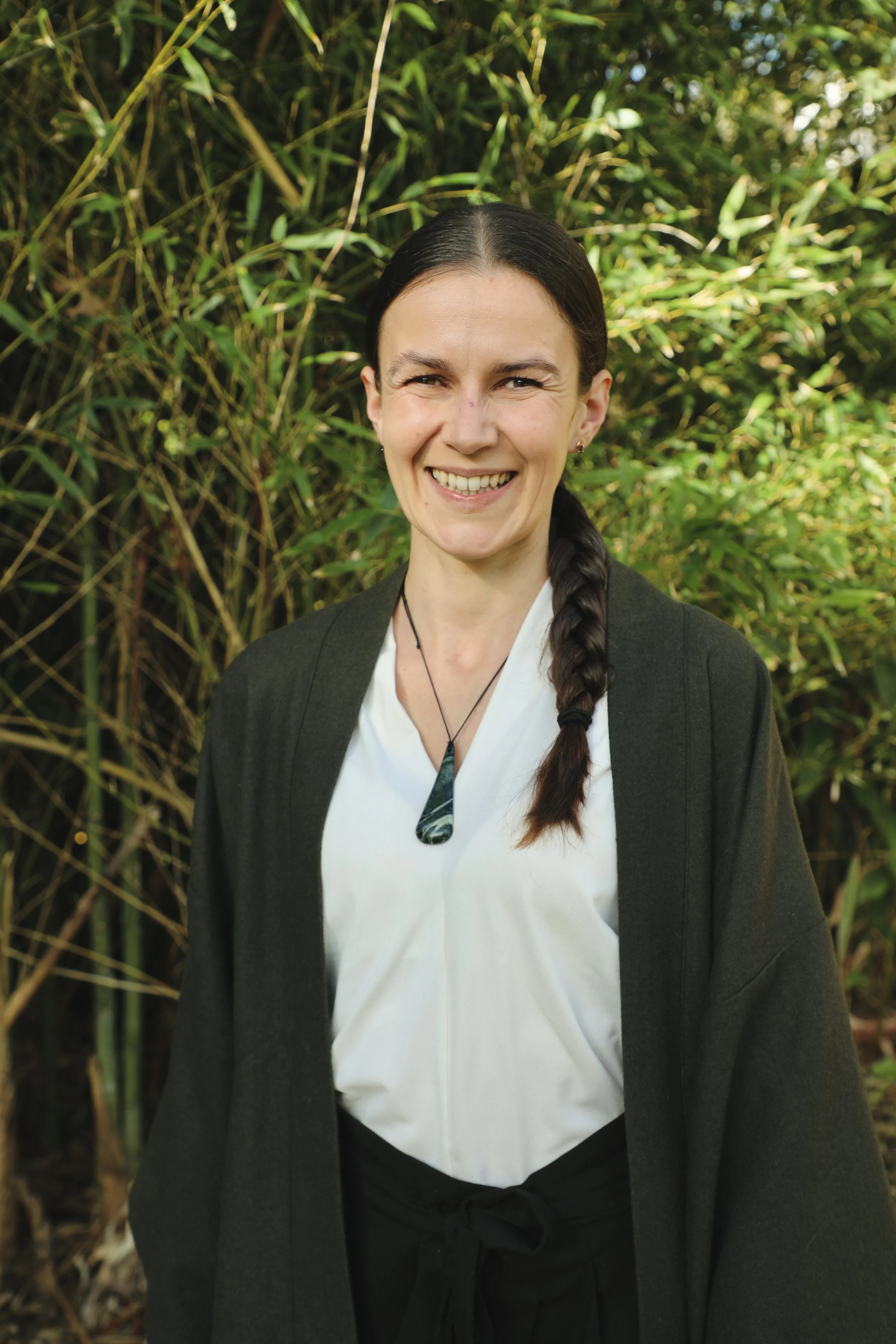 A woman with dark hair in a braid smiling outdoors, wearing a dark blazer, white blouse, and a necklace, with a background of green bamboo plants.