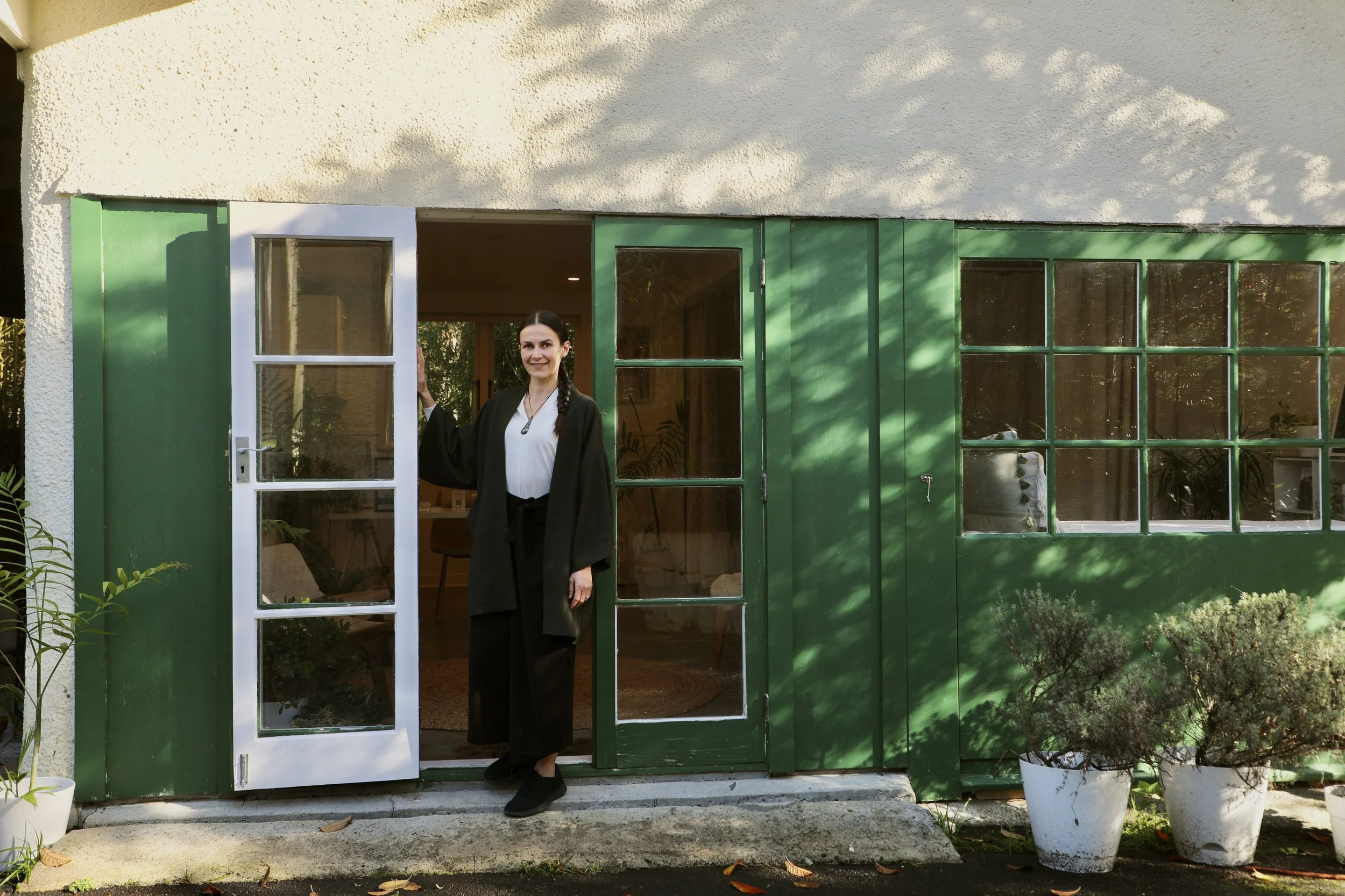 A woman standing in an open doorway of a house with green window frames, smiling at the camera. The house has a white stucco exterior and potted plants nearby.